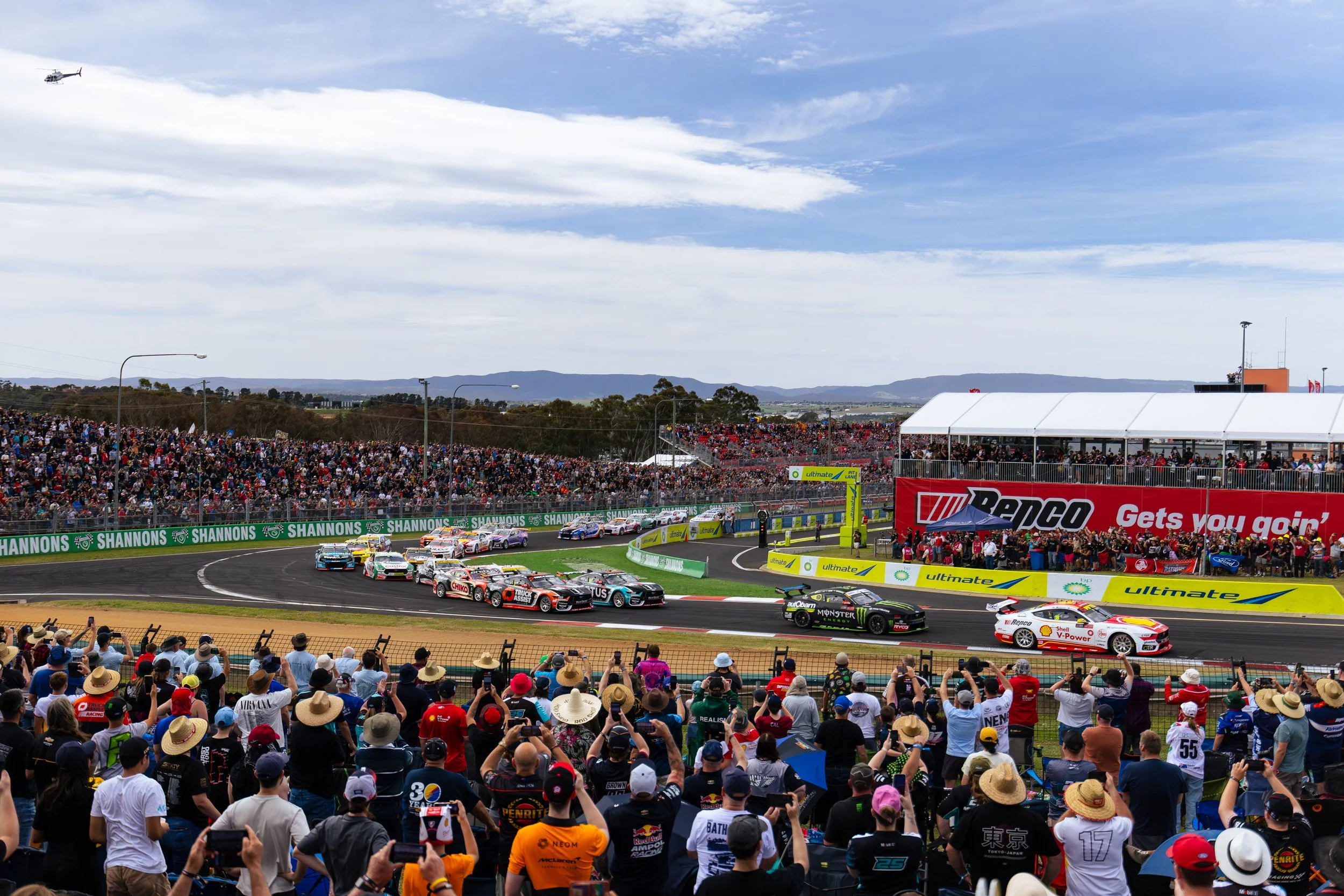 Start of the 2025 Bathurst 1000 viewed from the outside of turn 1 with the crowd in the foreground as the cars roar past.