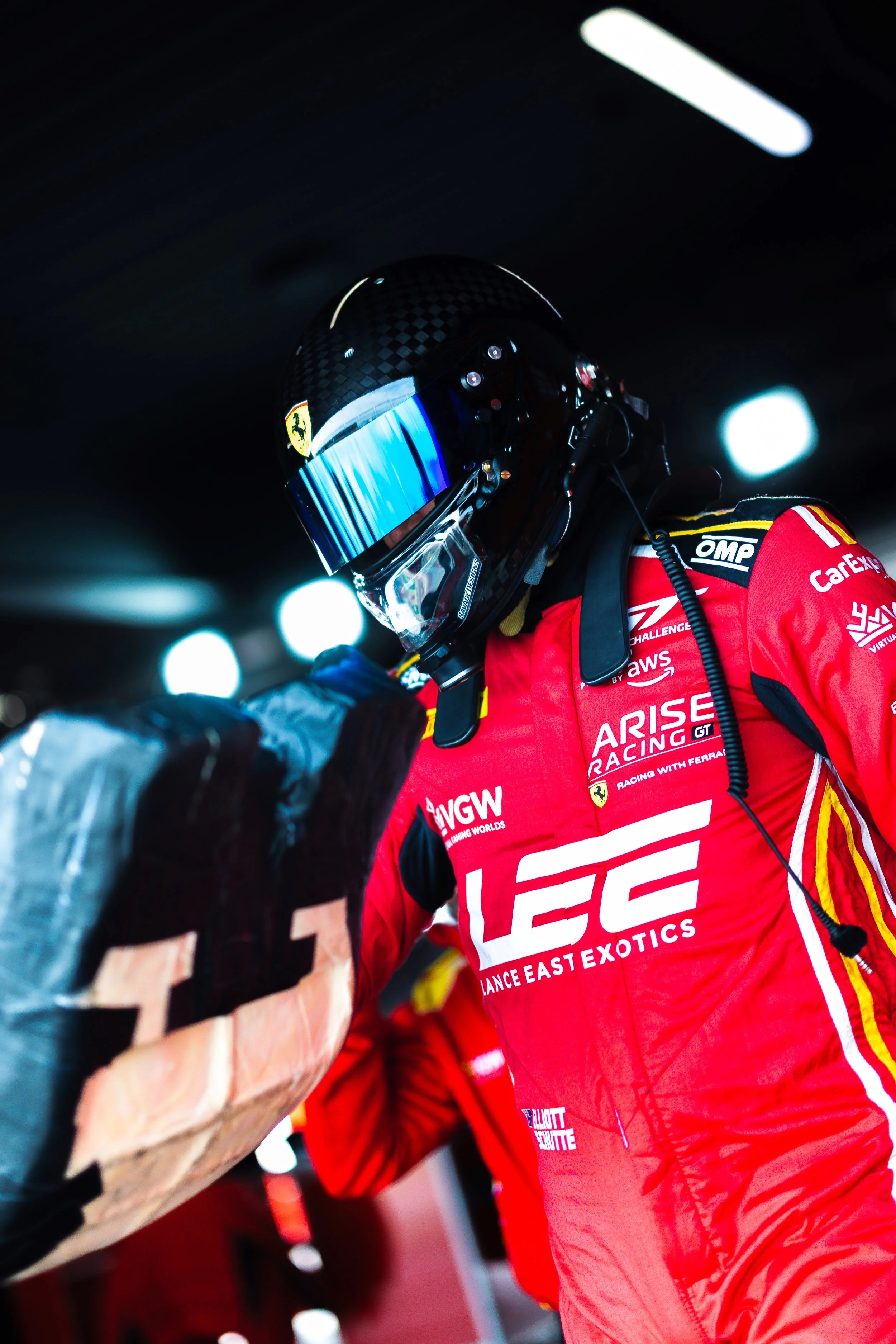 Elliot Schutte holding his seat insert with visor down, walking toward the Arise Racing GT3 Ferrari 296 during the 2025 GT World Challenge Australia season.