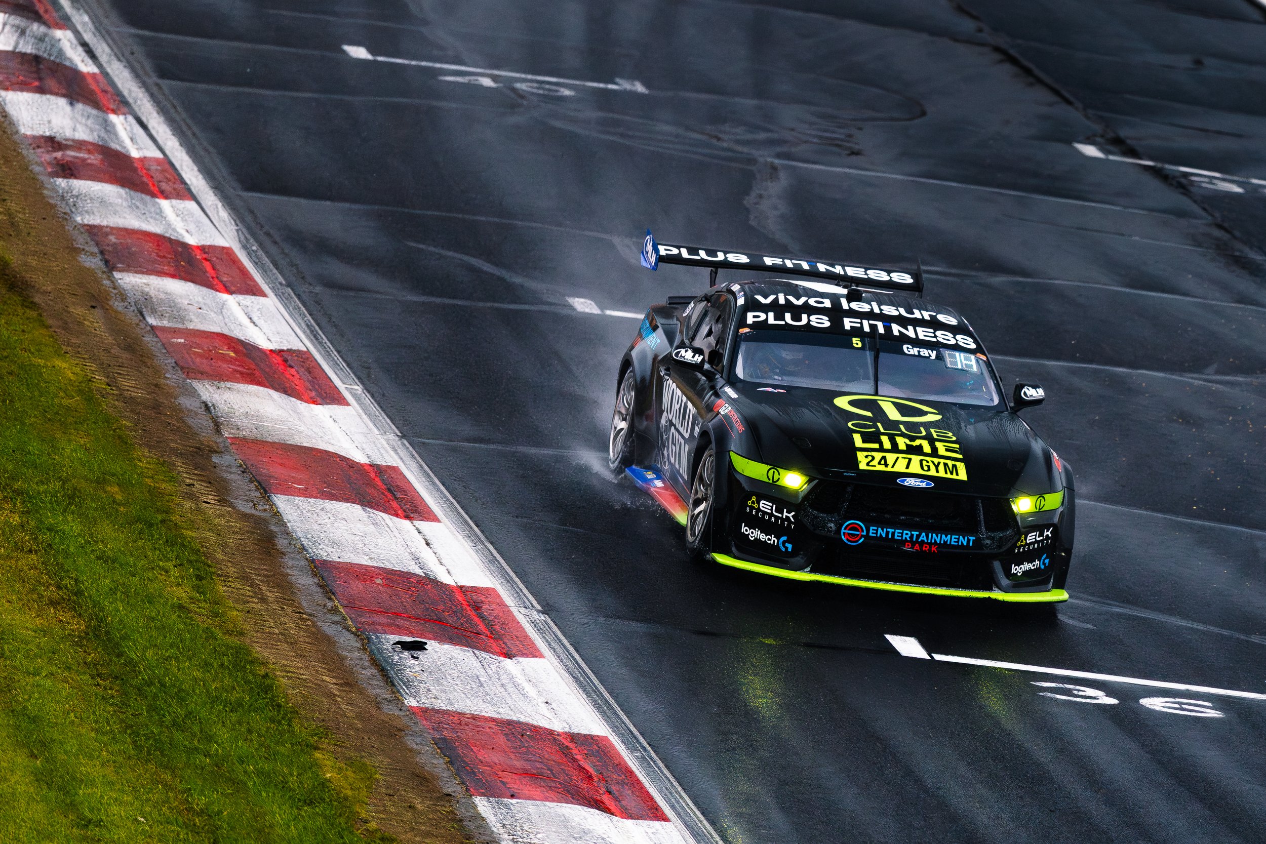 A black race car with bright yellow accents on a wet race track, kicking up spray as it takes a turn near the curb marked with red and white stripes.