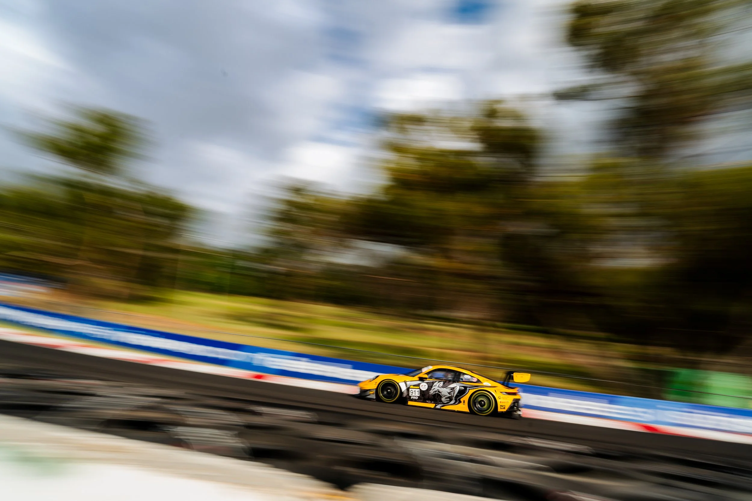 A yellow race car speeding on a racetrack with trees and sky in the background, with motion blur.