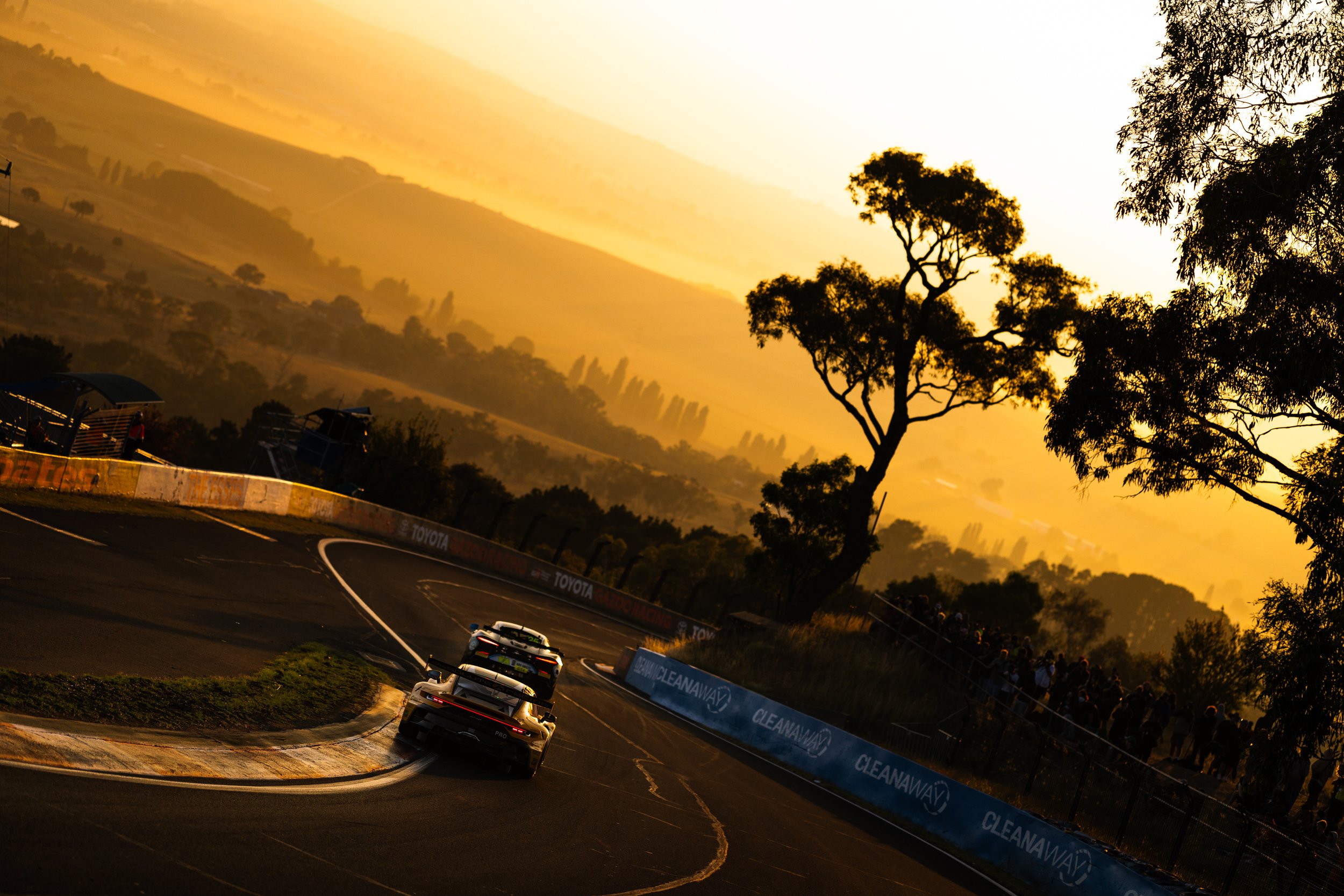 Race cars on a track during sunset with a mountain landscape in the background and spectators watching from the side.