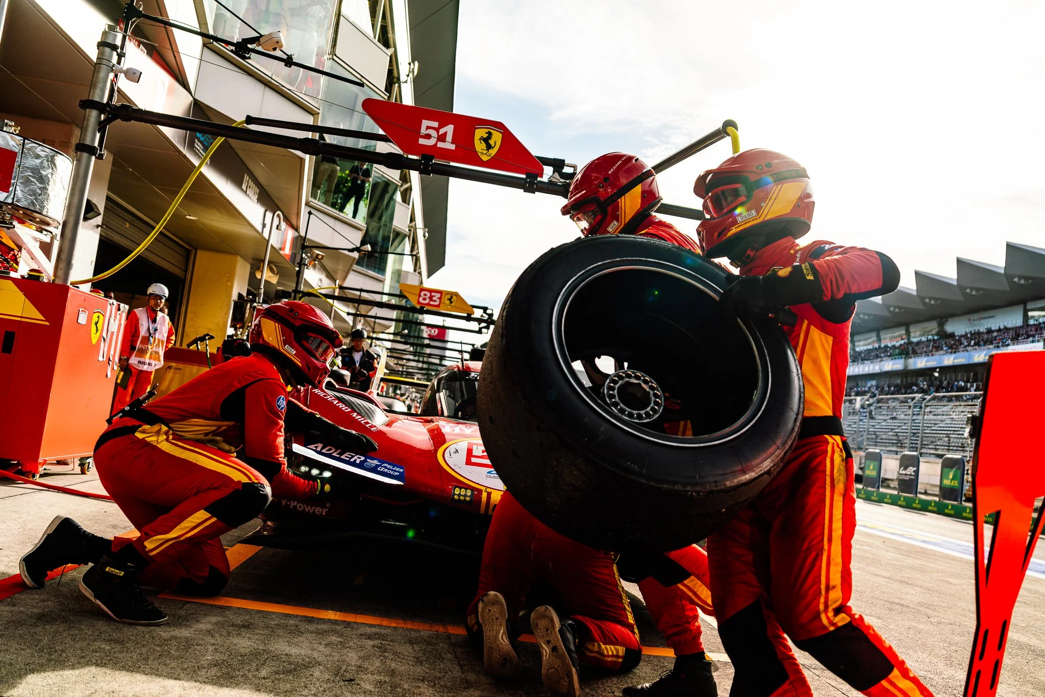 A Ferrari 499p race car is serviced by the team in the pit lane as a mechanic runs through the frame holding a tyre at the WEC Fuji Speedway round 2024.