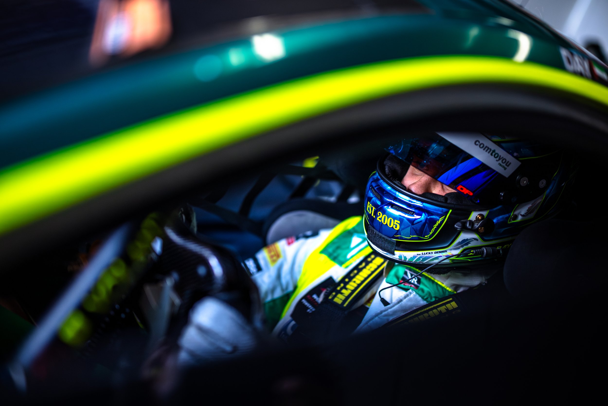 A race car driver wearing a blue helmet with yellow lettering, sitting inside a race car, with the door frame and interior visible.