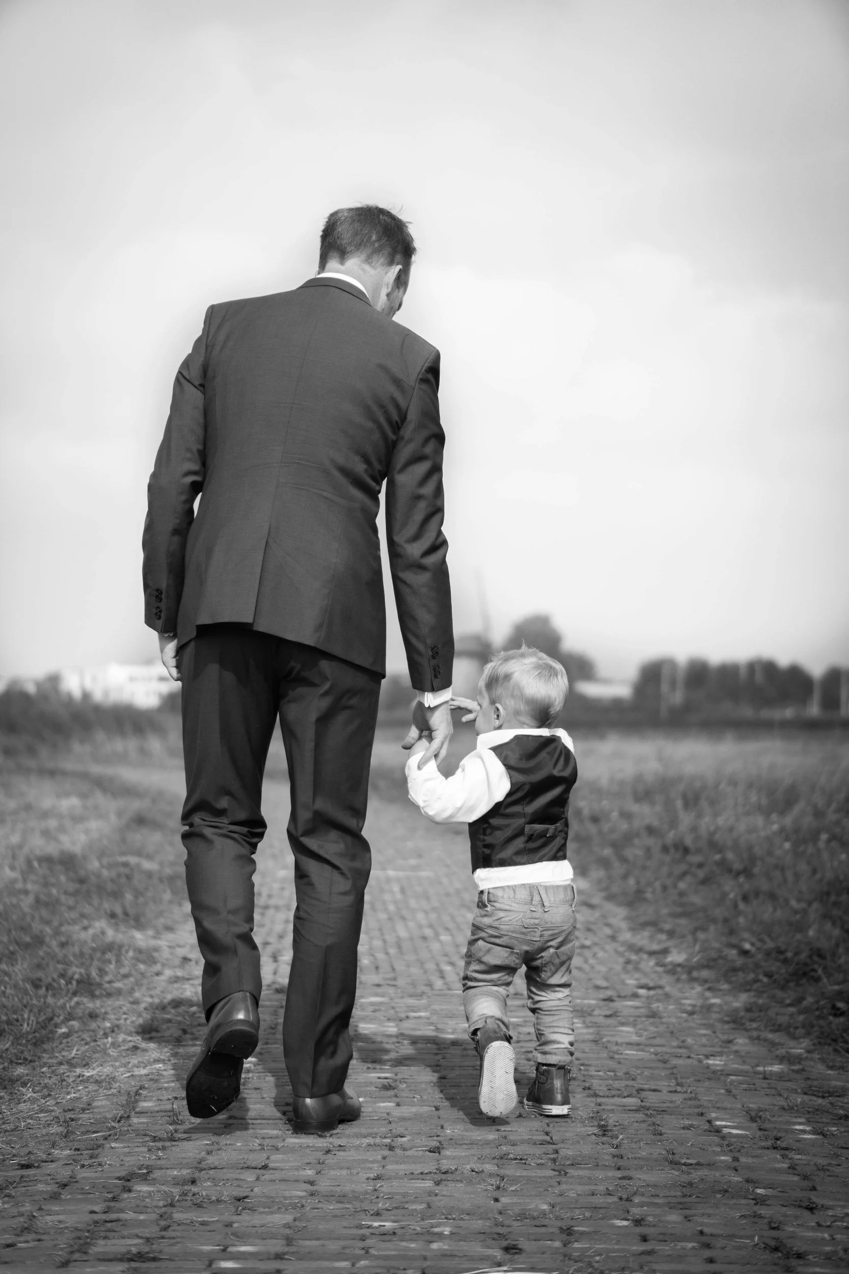 A man in a suit holding the hand of a young boy walking on a cobblestone path outdoors.