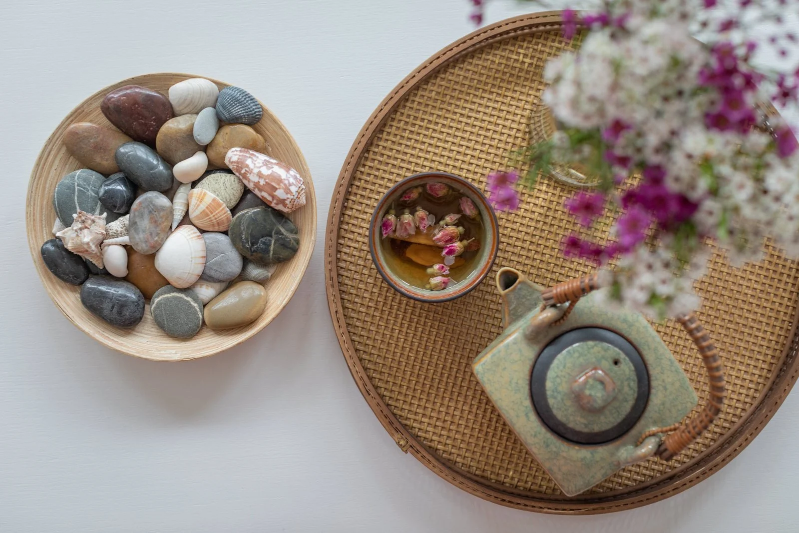 A white table with a bamboo tray holding a ceramic teapot and a mug with dried flowers, next to a round woven tray, and a separate bowl filled with smooth, polished stones and seashells, with a vase of pink and white flowers on the tray.