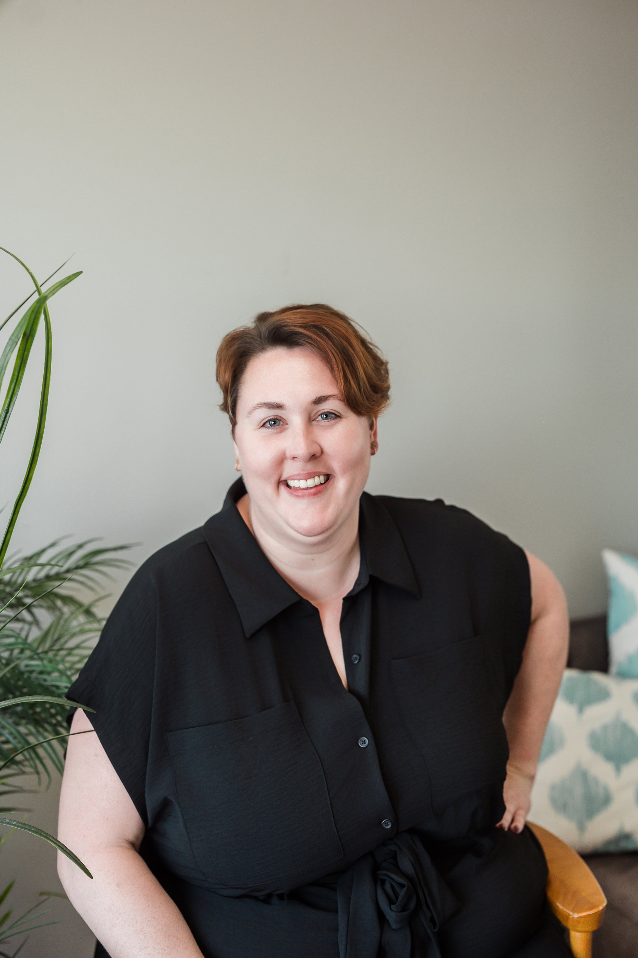 A woman with short reddish-brown hair, wearing a black sleeveless button-up shirt, sitting on a wooden chair in a room with light gray walls and a green potted plant nearby, smiling at the camera.