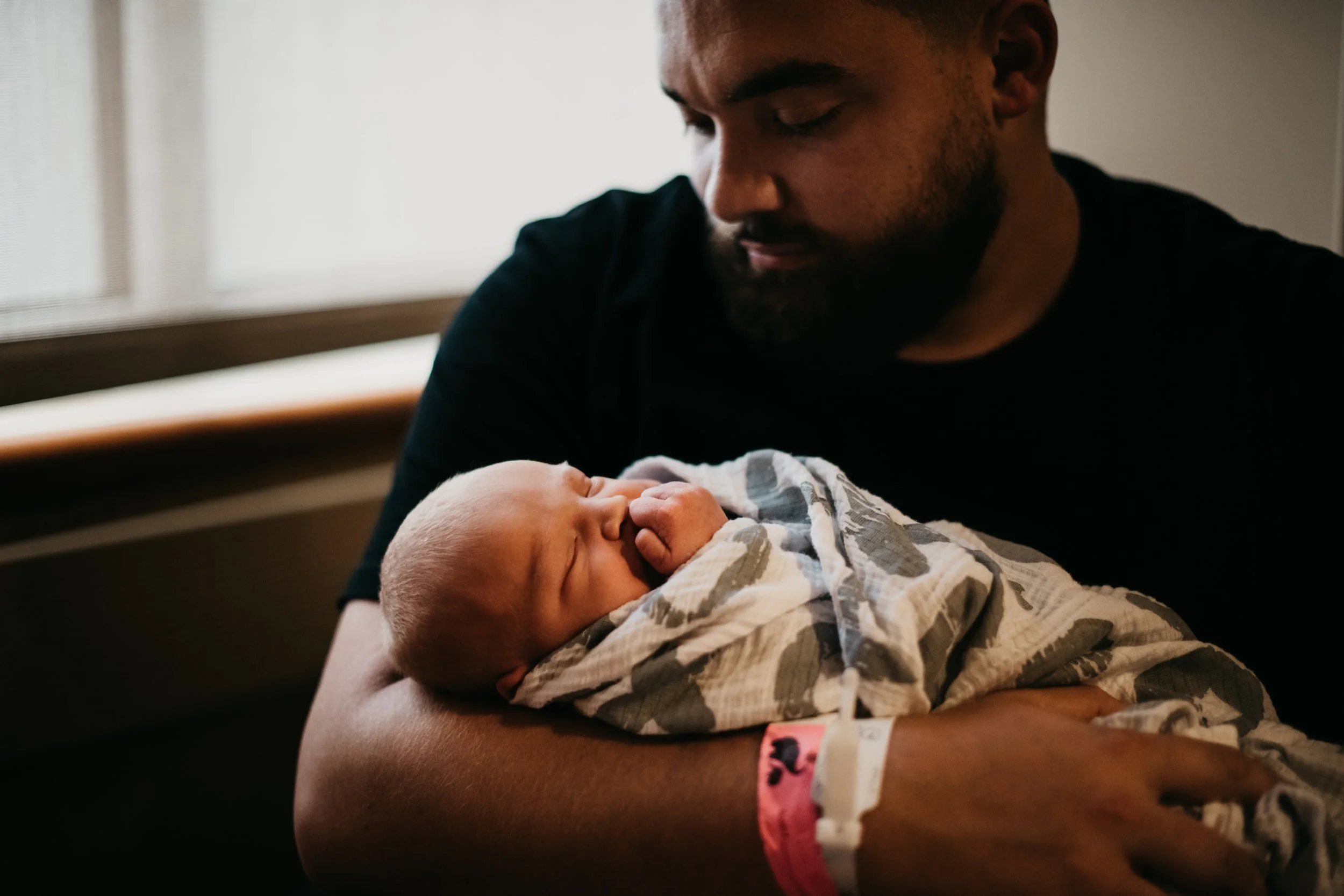 African American man sitting in front of a window holding a newborn baby wrapped in a blanket or swaddle