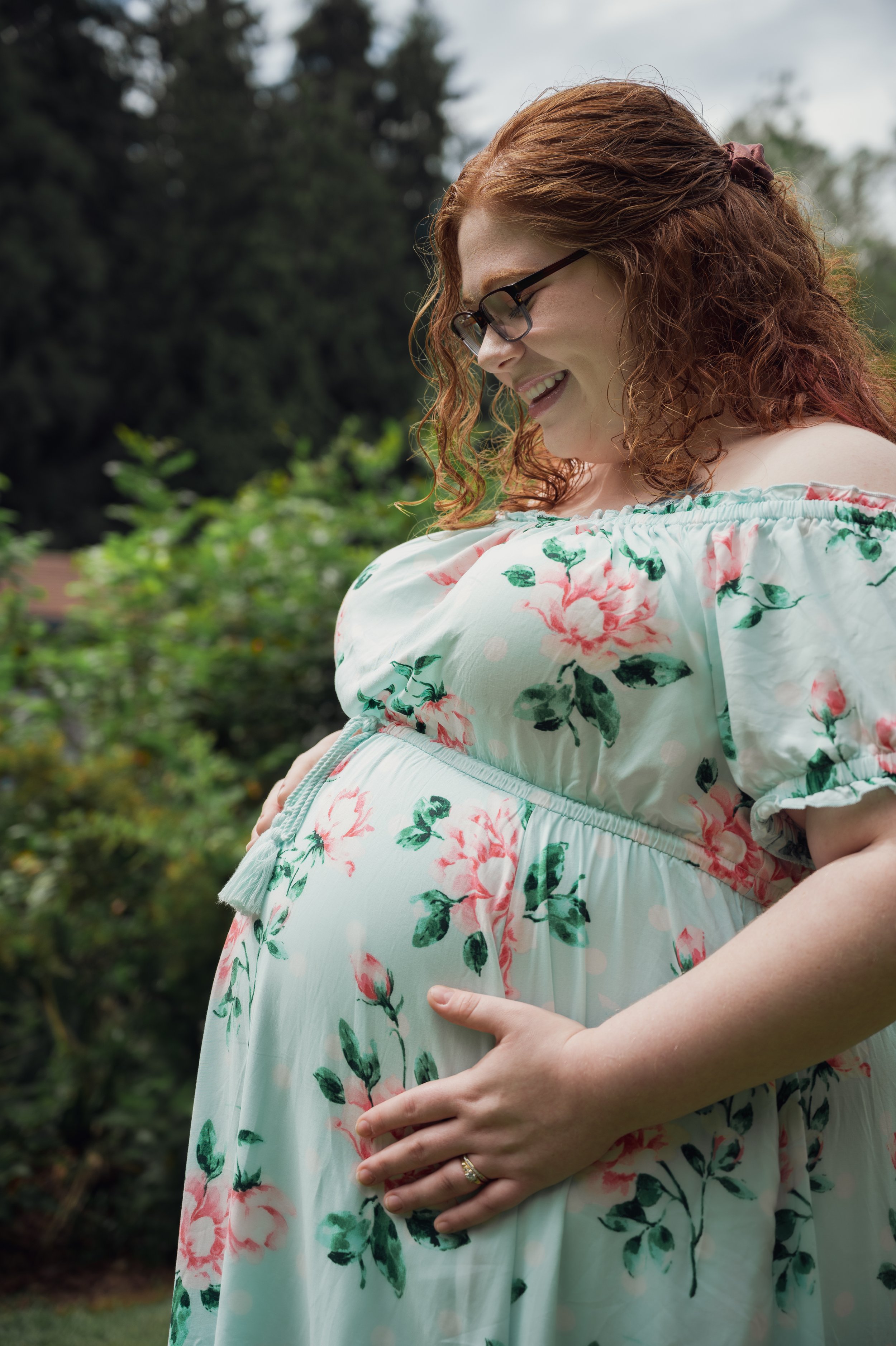 A pregnant woman with long curly red hair and glasses wearing a white dress with a pink and green floral pattern, standing outdoors, smiling with her hands on her baby bump.
