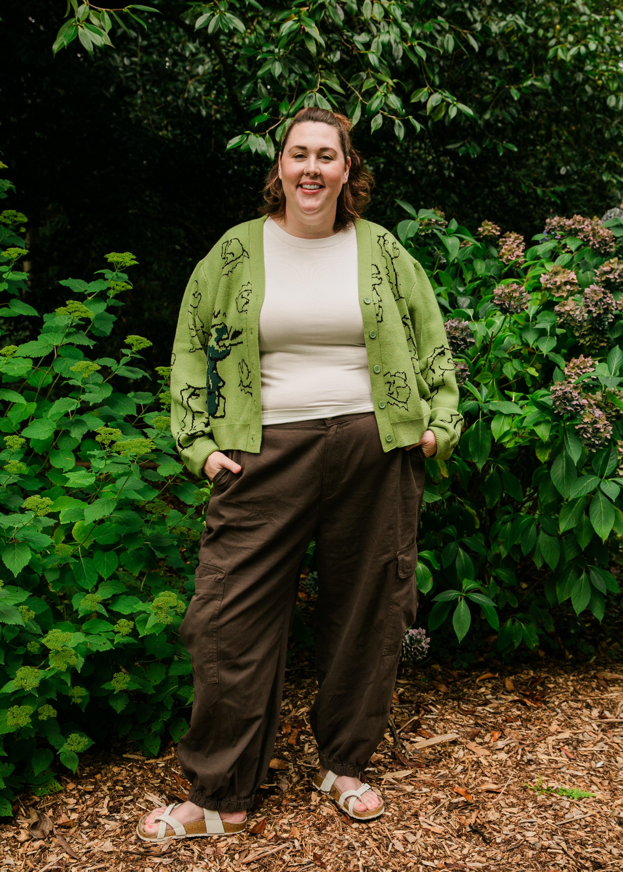 A woman standing outdoors in a garden with green foliage and pink hydrangea flowers, wearing a beige t-shirt, a green cardigan with black line drawings, brown cargo pants, and white sandals.