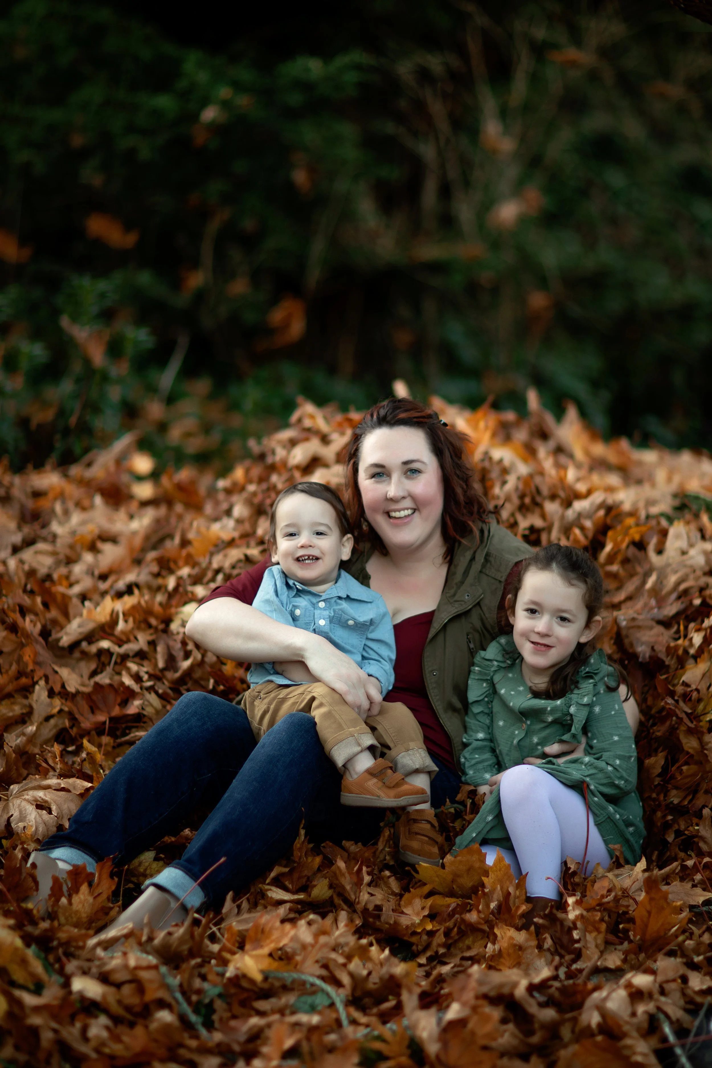 A woman with two children sitting on a bed of fallen autumn leaves outdoors, with trees and bushes in the background.