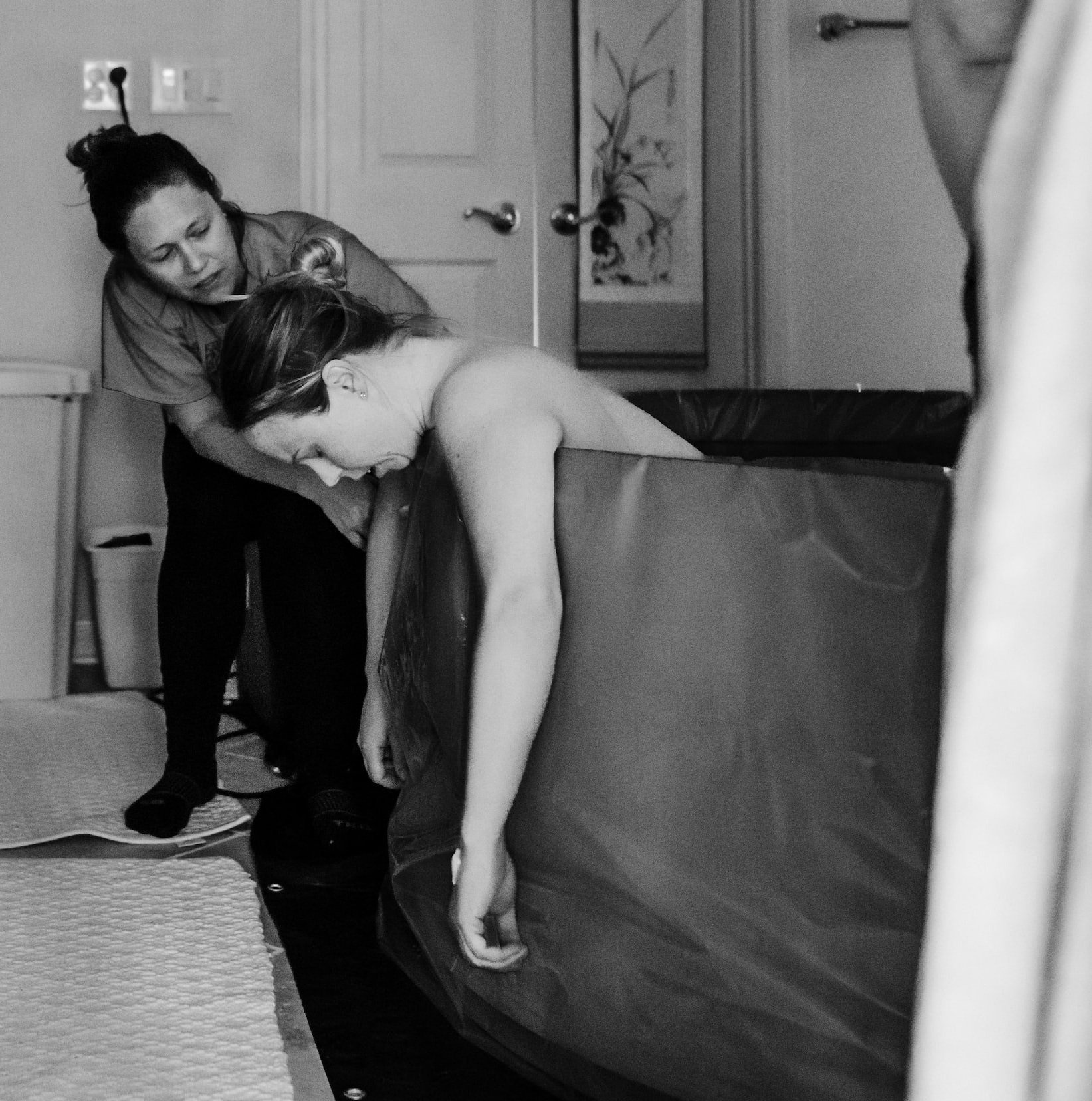 woman in an at home birthing tub, leaning over the side, supported by a doula standing over her leaning down to help her