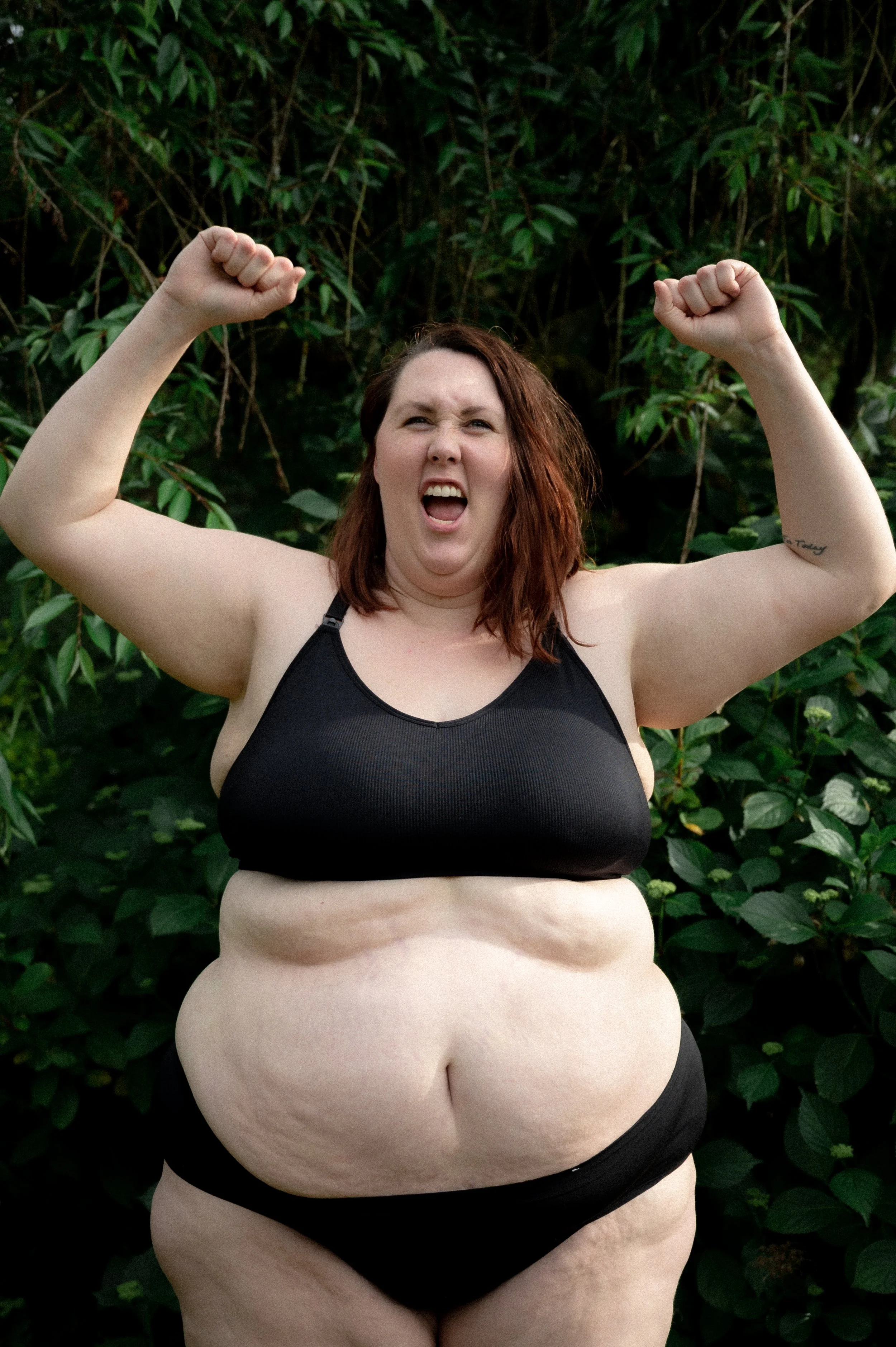 A woman with red hair and fair skin, wearing black sports bra and black underwear, flexing her arms with a determined expression, in front of green foliage.
