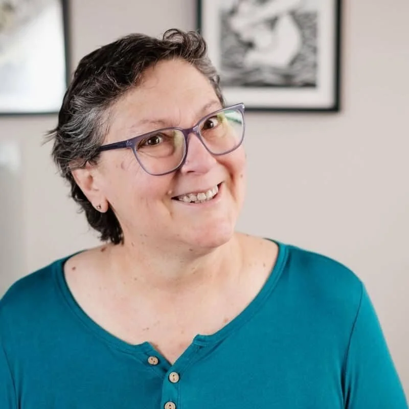 A woman with short gray hair, glasses, and a teal shirt smiling in a room with framed art on the wall.