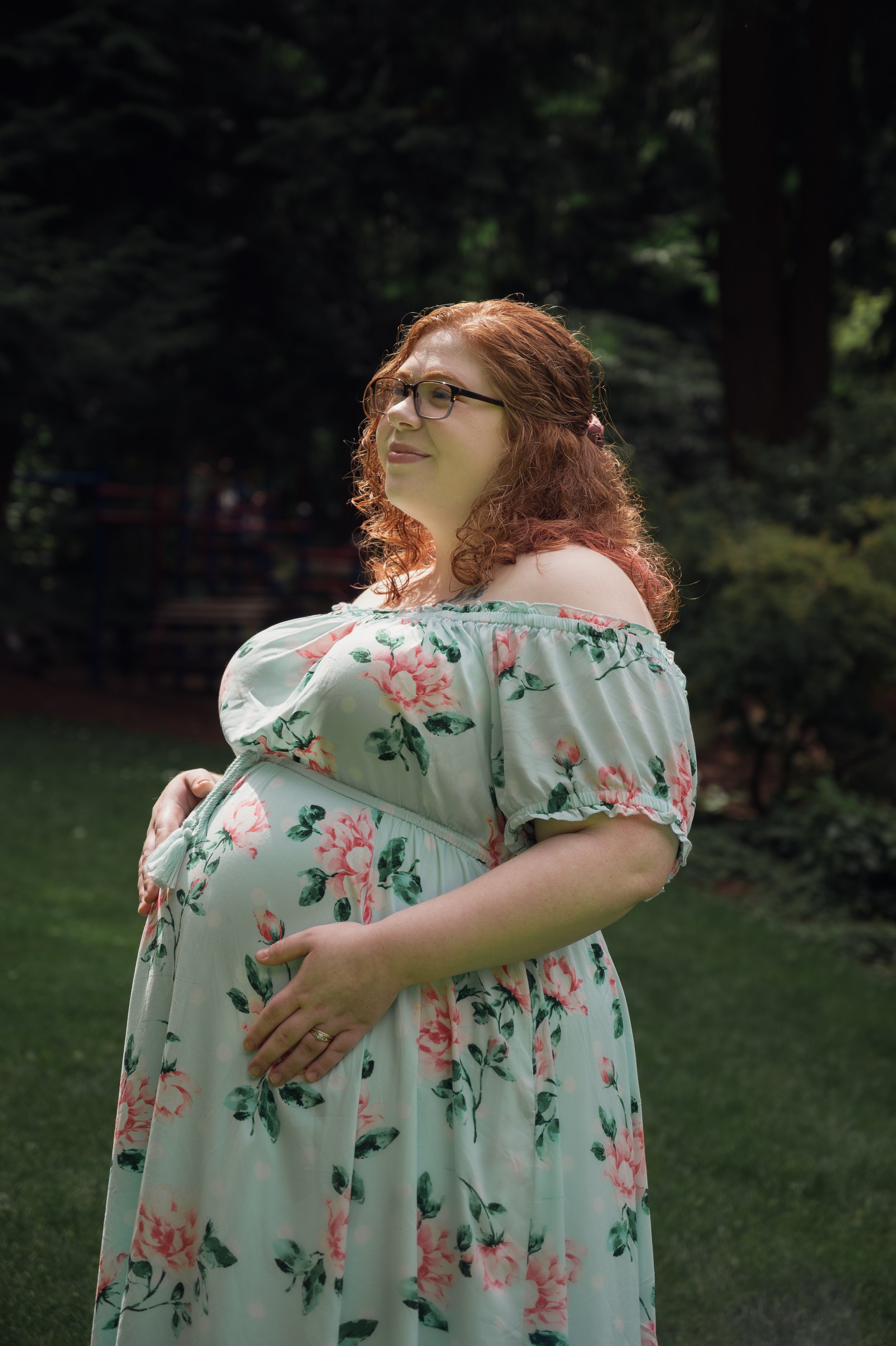 red headed woman with glasses in a flower dress in a sideview image, the pregnant woman has her hands on her pregnant belly