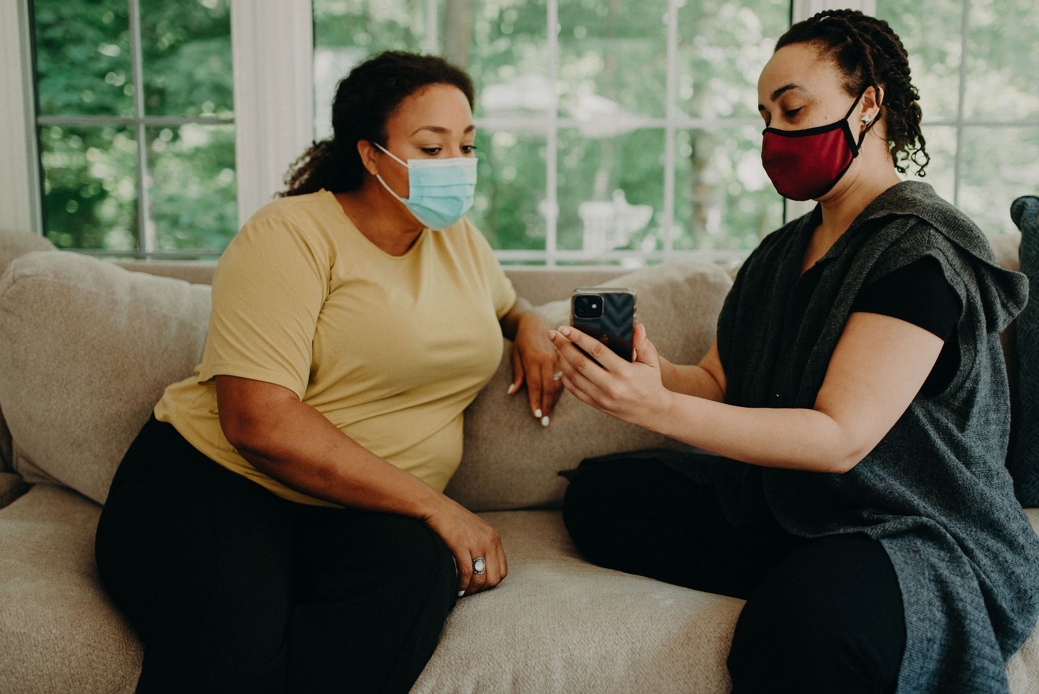 Two women sitting on a sofa, both wearing face masks and looking at a smartphone together.