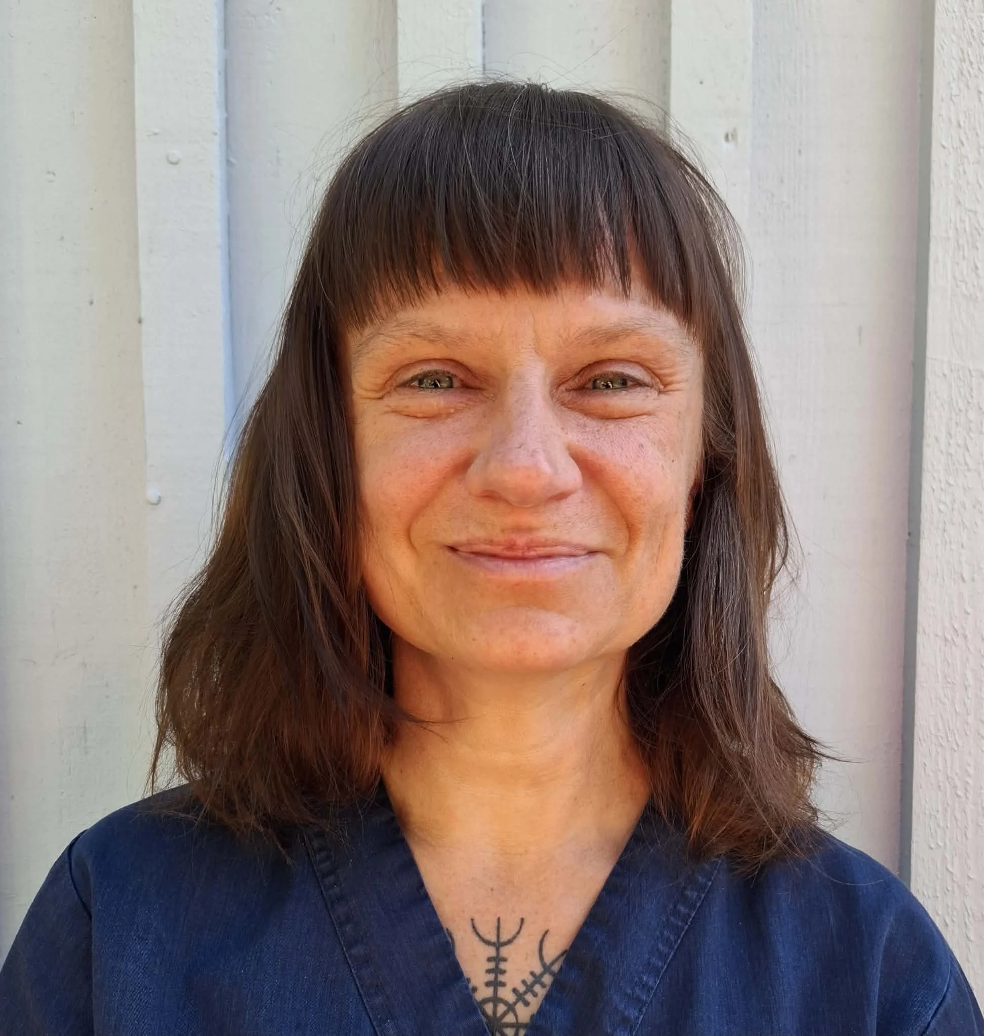 En kvinna med mörka, raka hår och en tatuering i halsen, som ler och bär en mörkblå tröja, står framför en vit vägg.