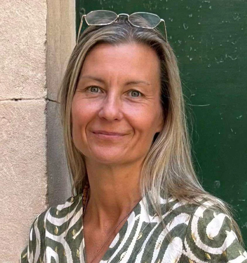Kvinna med långt blont hår, solglasögon på huvudet, står framför en grön dörr och en vägg, ler lätt.