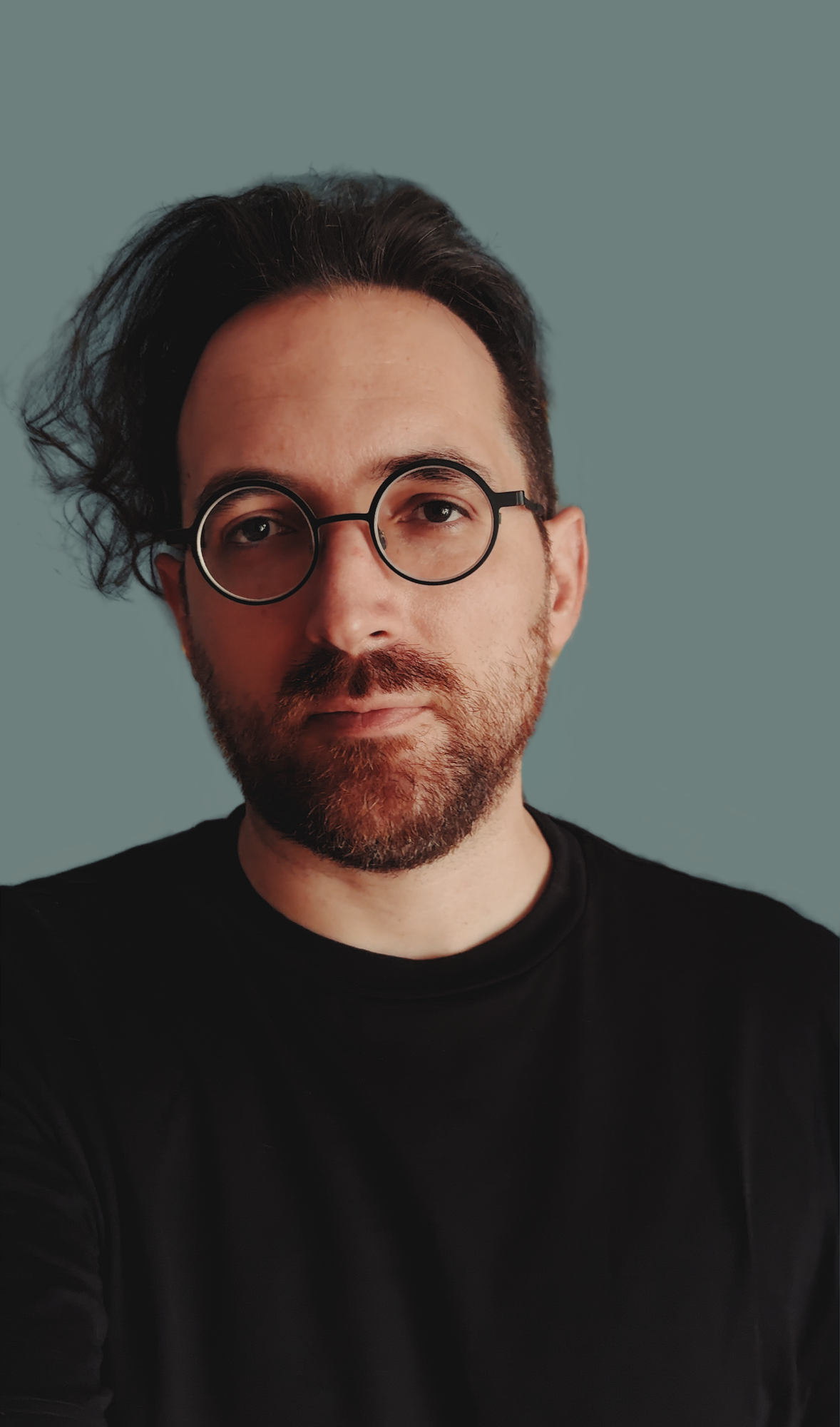A man with dark, messy hair, a beard, wearing round glasses and a black shirt, standing against a gray background.