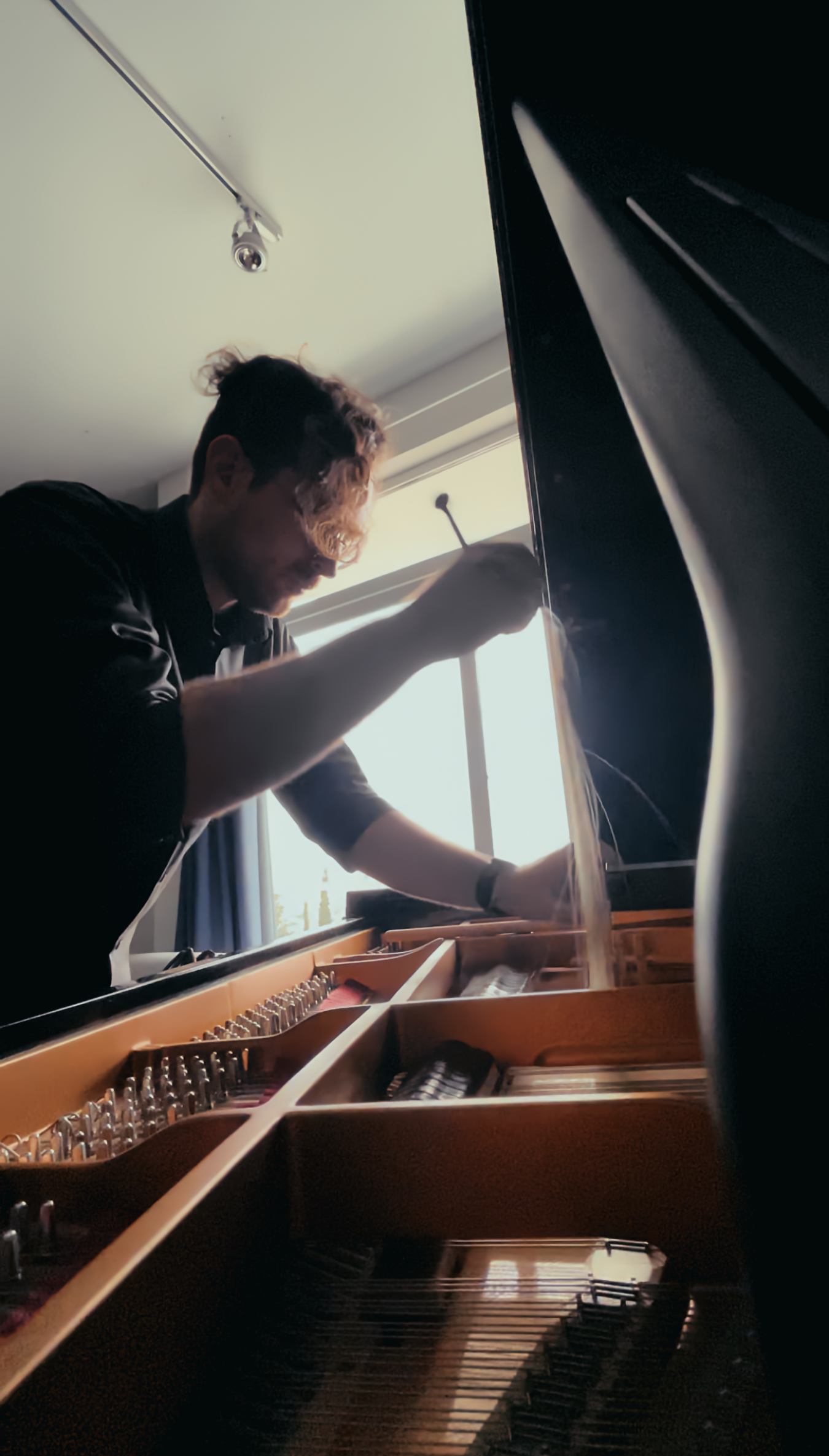 A person working on a grand piano, seen from inside the piano, with sunlight coming through a window in the background.