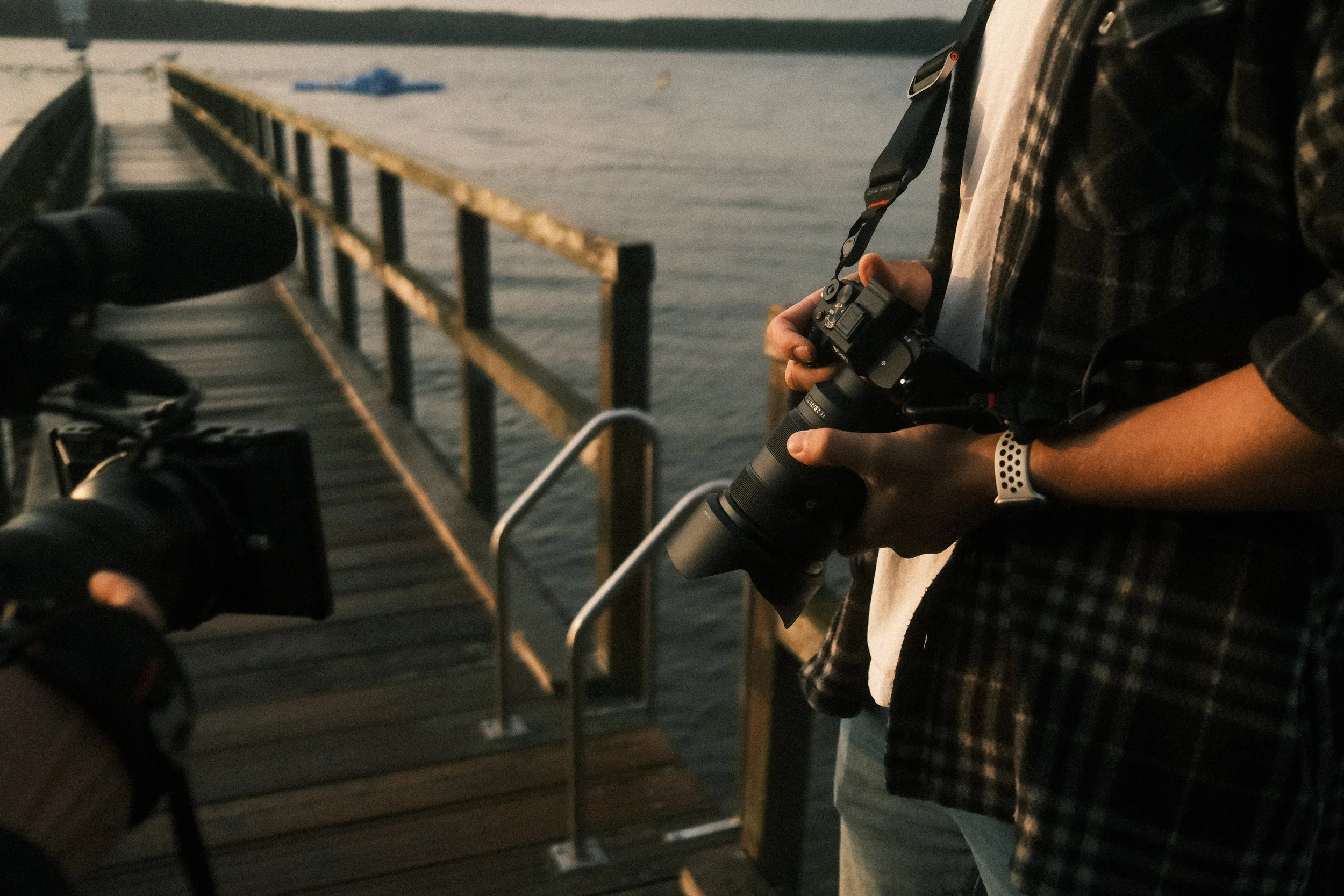 Person hält eine Kamera auf einem Holzsteg am Wasser während eines fotografischen Moments im warmen Abendlicht
