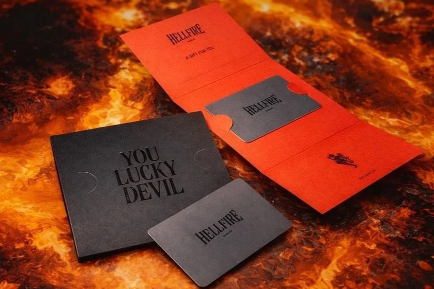 A black card case with the words "YOU LUCKY DEVIL" on it, and a red bookmark with the word "HELLFIRE" on it, sitting on a fiery patterned surface.