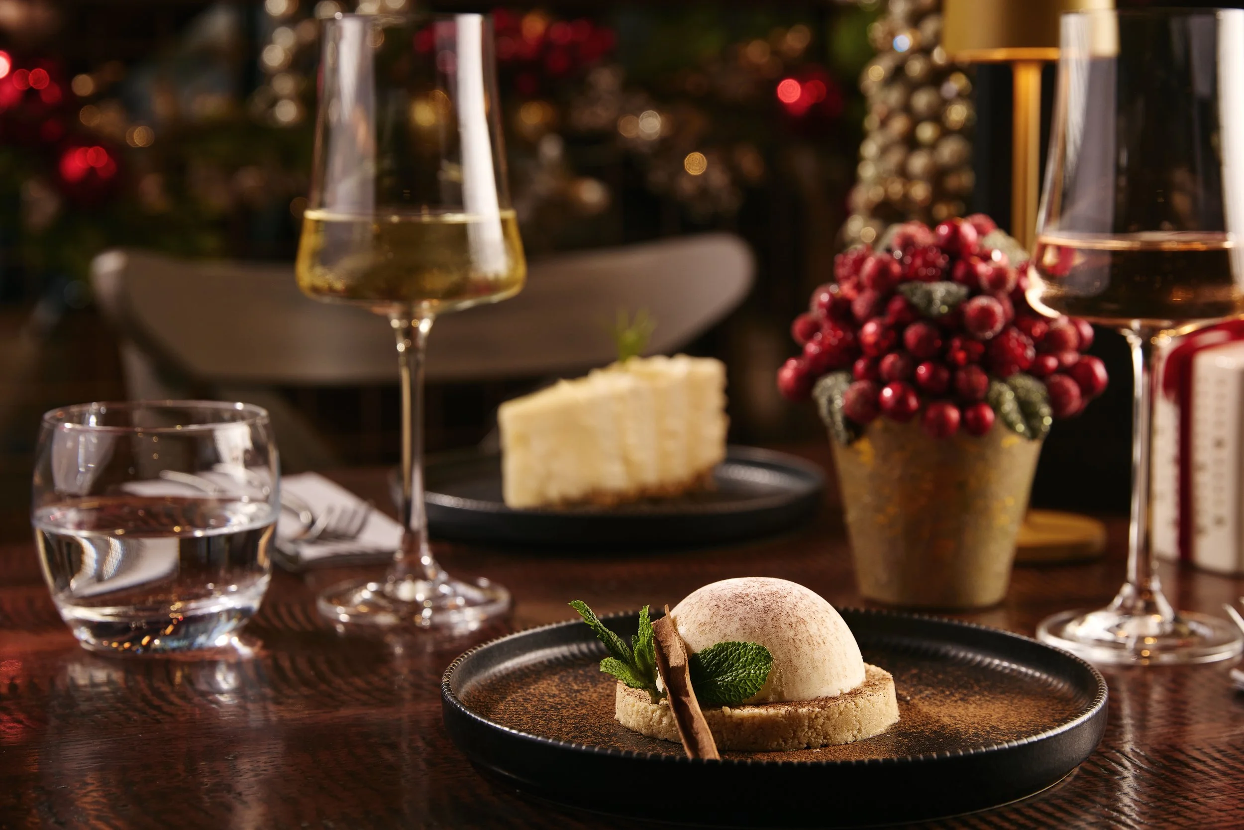 A festive dinner table with two glasses of champagne, a glass of water, a slice of cheesecake, a dessert with mint and chocolate, and a potted plant with red berries, set for a holiday celebration.