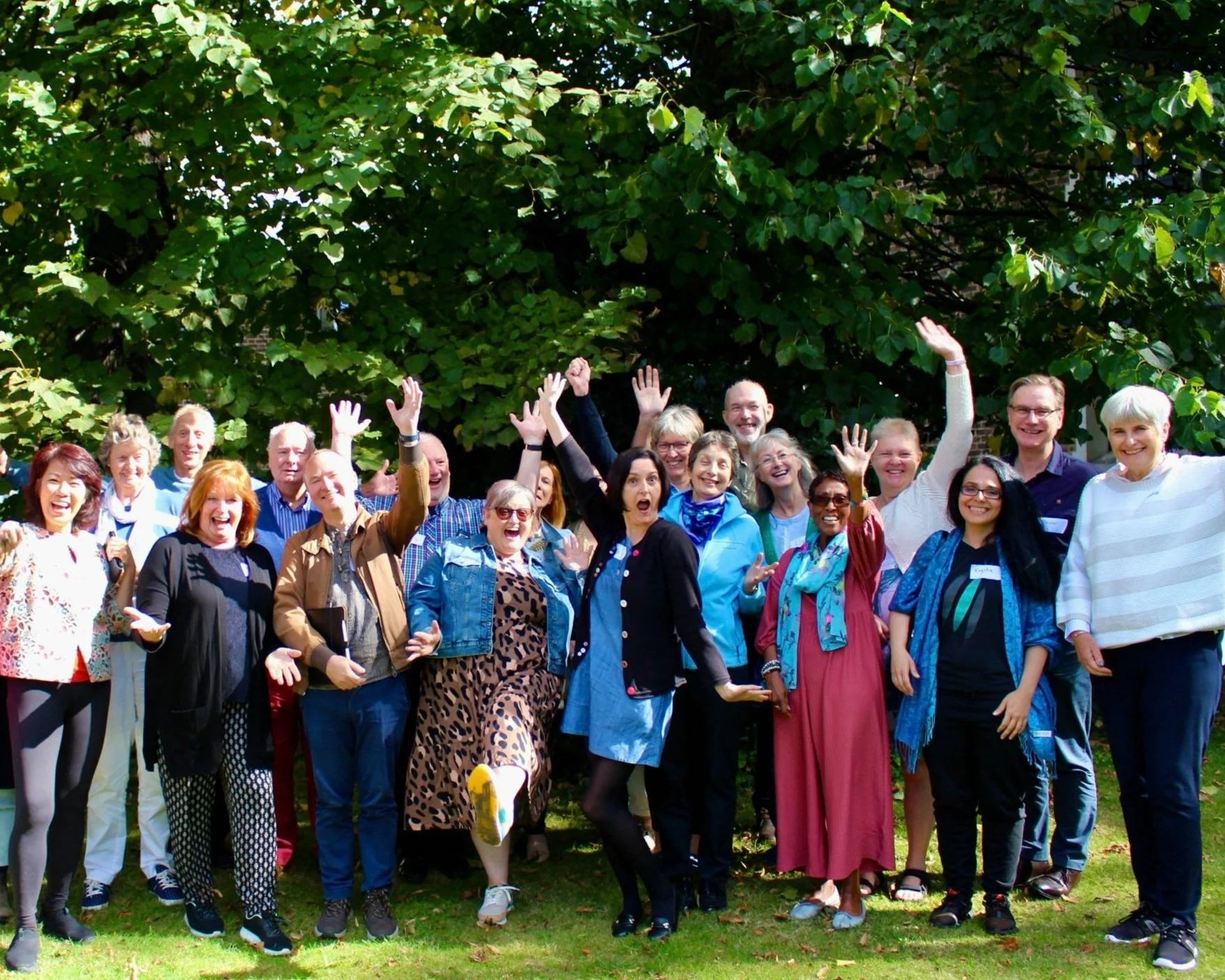 Group of people smiling and posing outdoors under a tree, all with raised hands, on a sunny day.