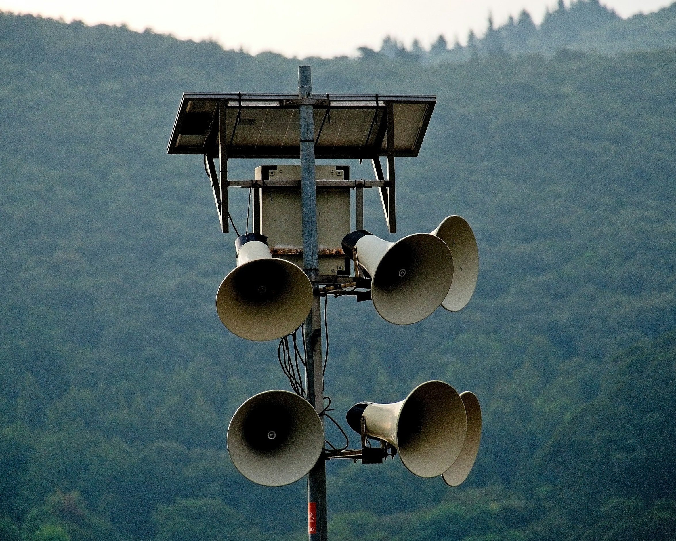 A pole with multiple loudspeakers and a solar panel, set against a backdrop of green hills.