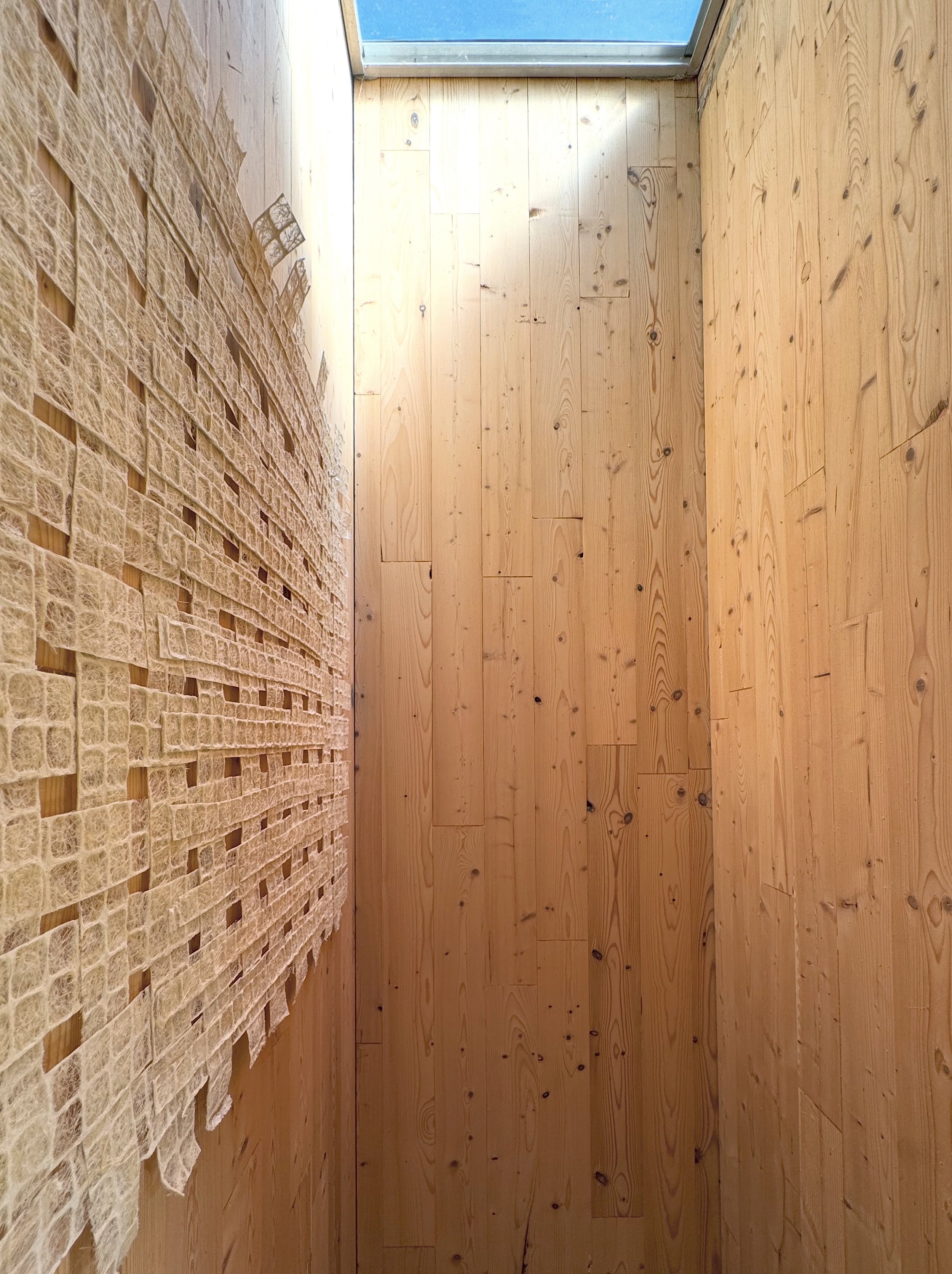 Wooden floor and wall, partially covered by beige textured material on the left side, with a window or glass door at the top of the image.