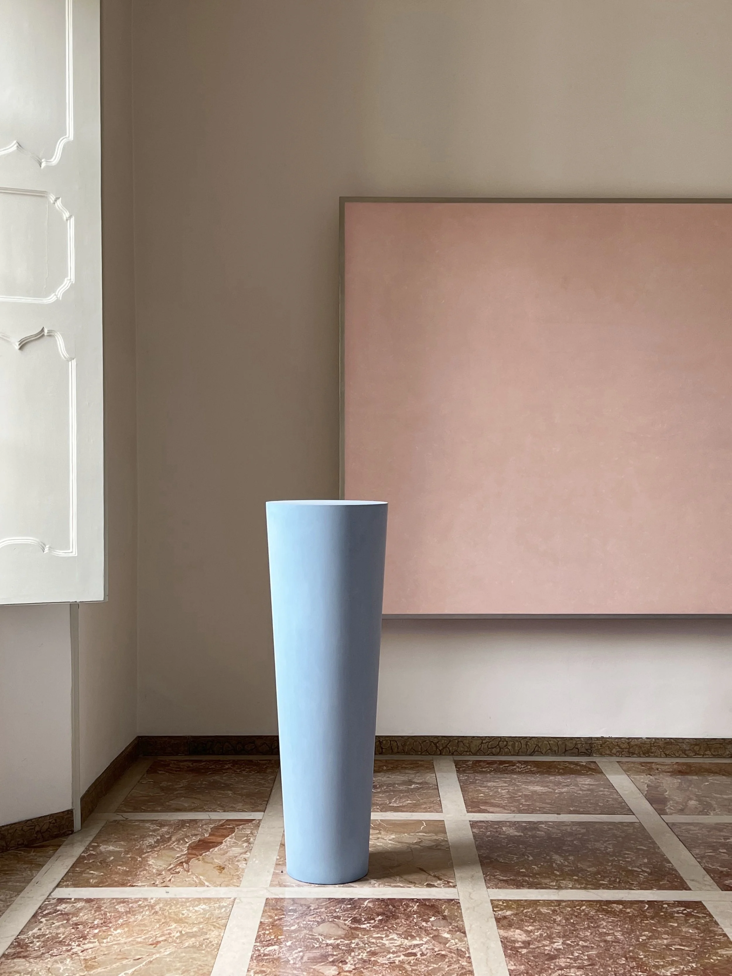 A tall, cylindrical, light blue vase standing on a checkered marble floor in a room with beige walls and a pinkish wall panel.