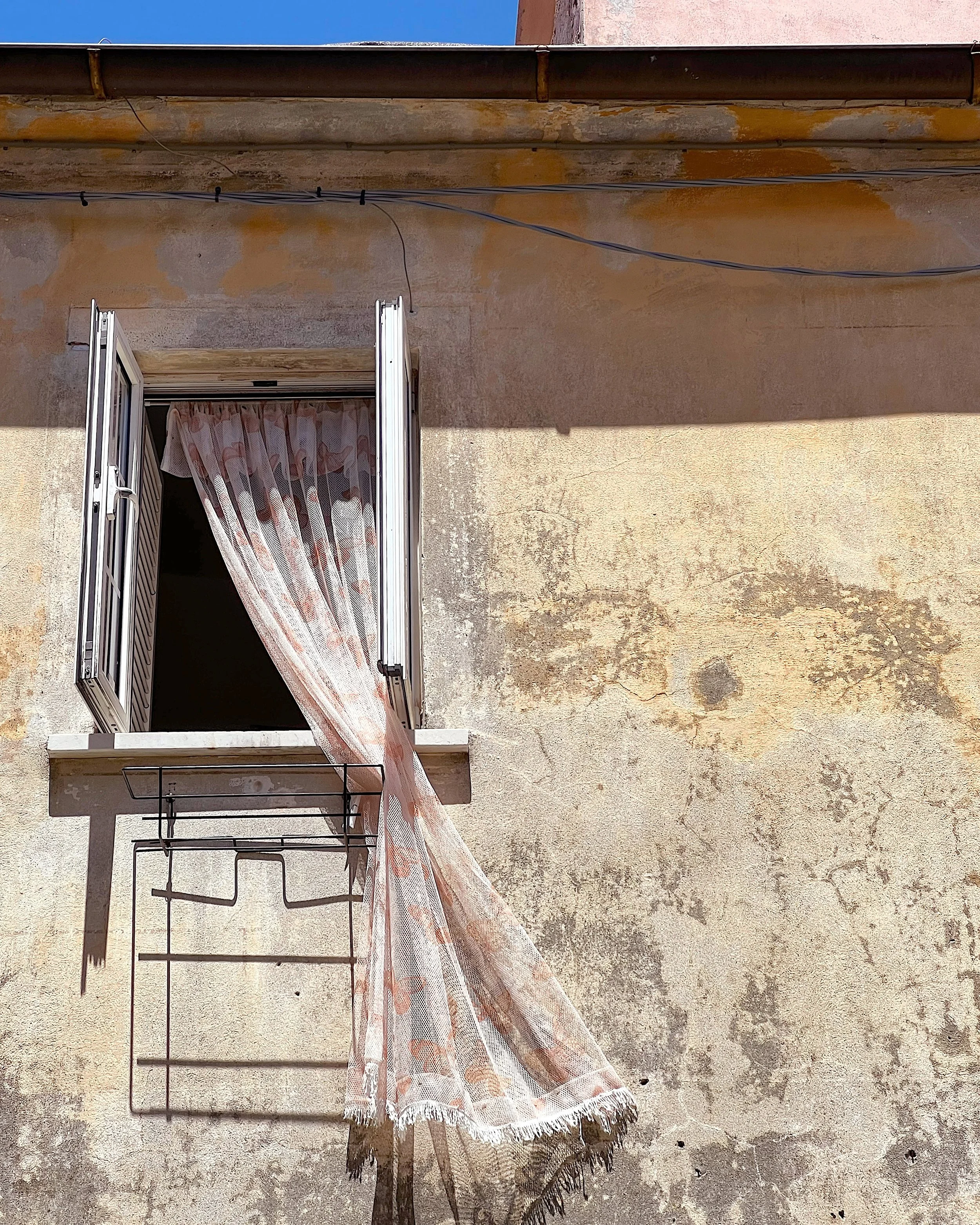 View of a rustic outdoor wall with a partially open window with floral curtains, a small black metal table, and a sheer peach-colored curtain hanging from the balcony.