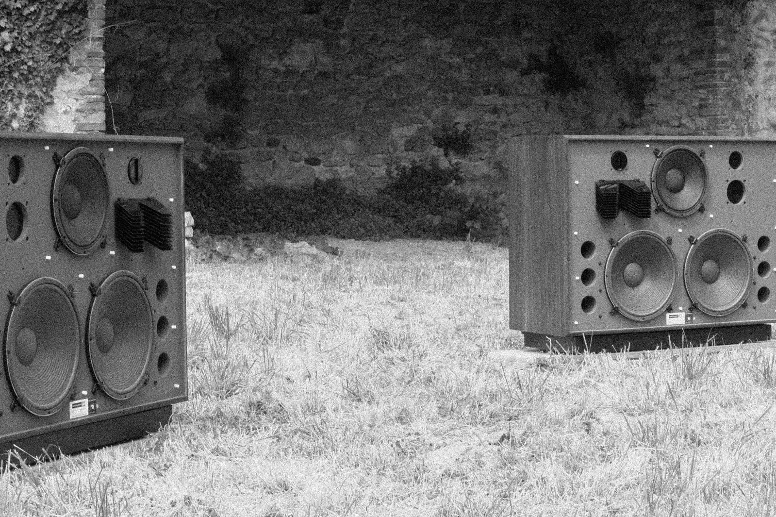 Two large speaker boxes placed outdoors on a grassy area with a stone wall and some bushes in the background.