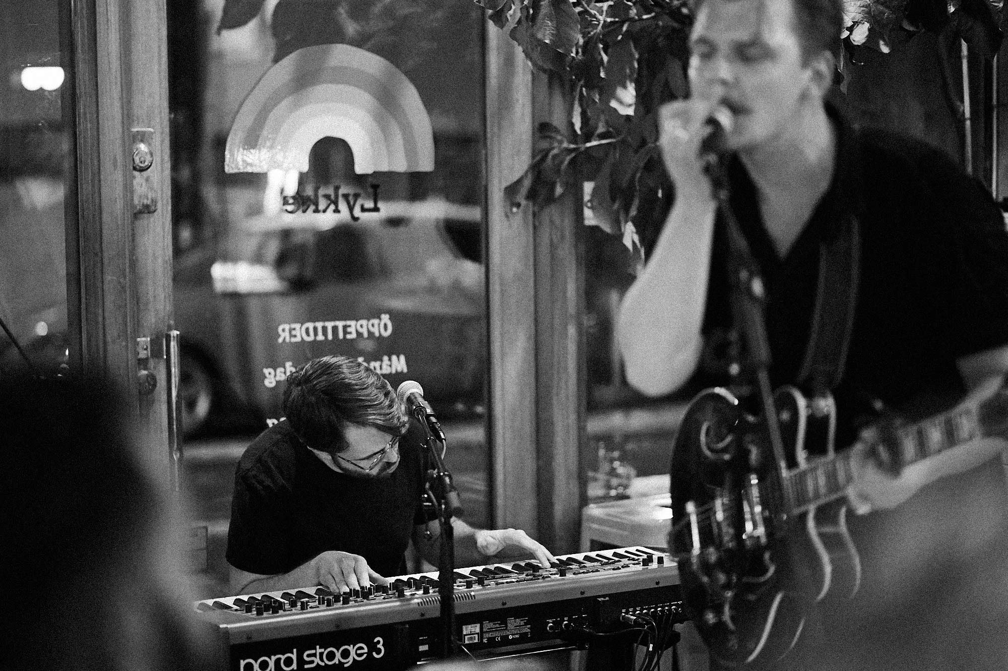 Two musicians perform in a dimly lit indoor space; one is on piano and the other on guitar, both focused on their instruments.