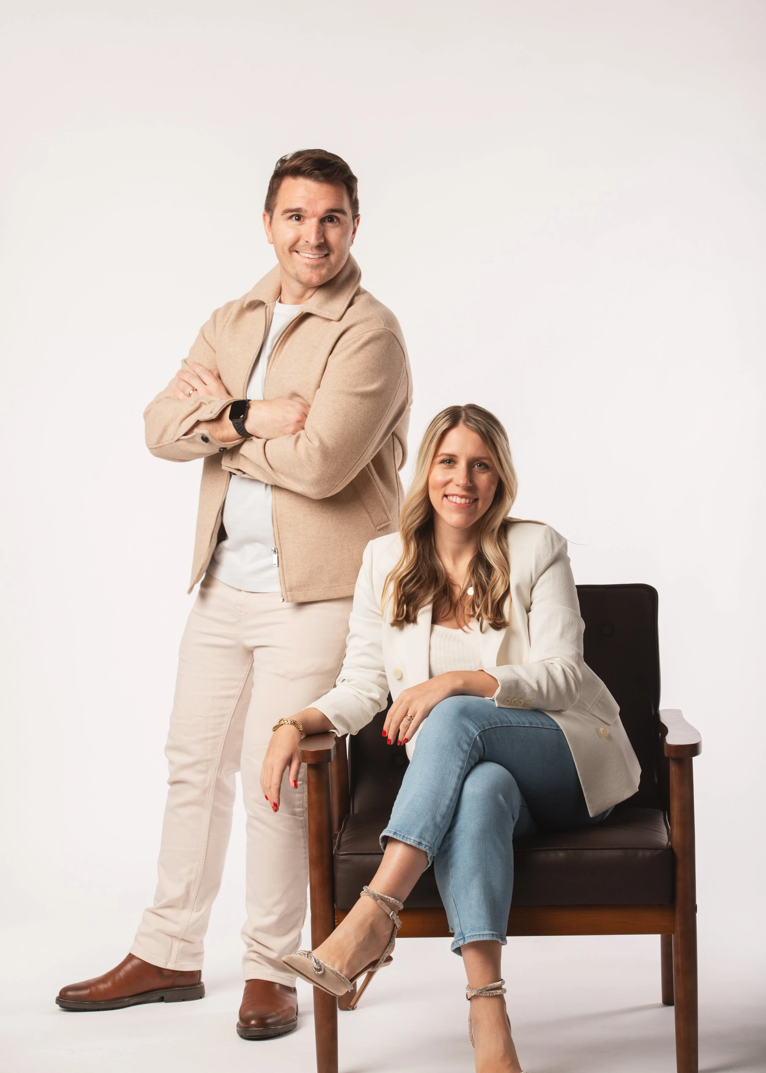 A man and a woman posing together against a plain white background. The man stands with arms crossed, wearing a beige jacket, white shirt, and light-colored pants. The woman sits in a chair, wearing a white blazer, light blue jeans, and beige heels, smiling at the camera.
