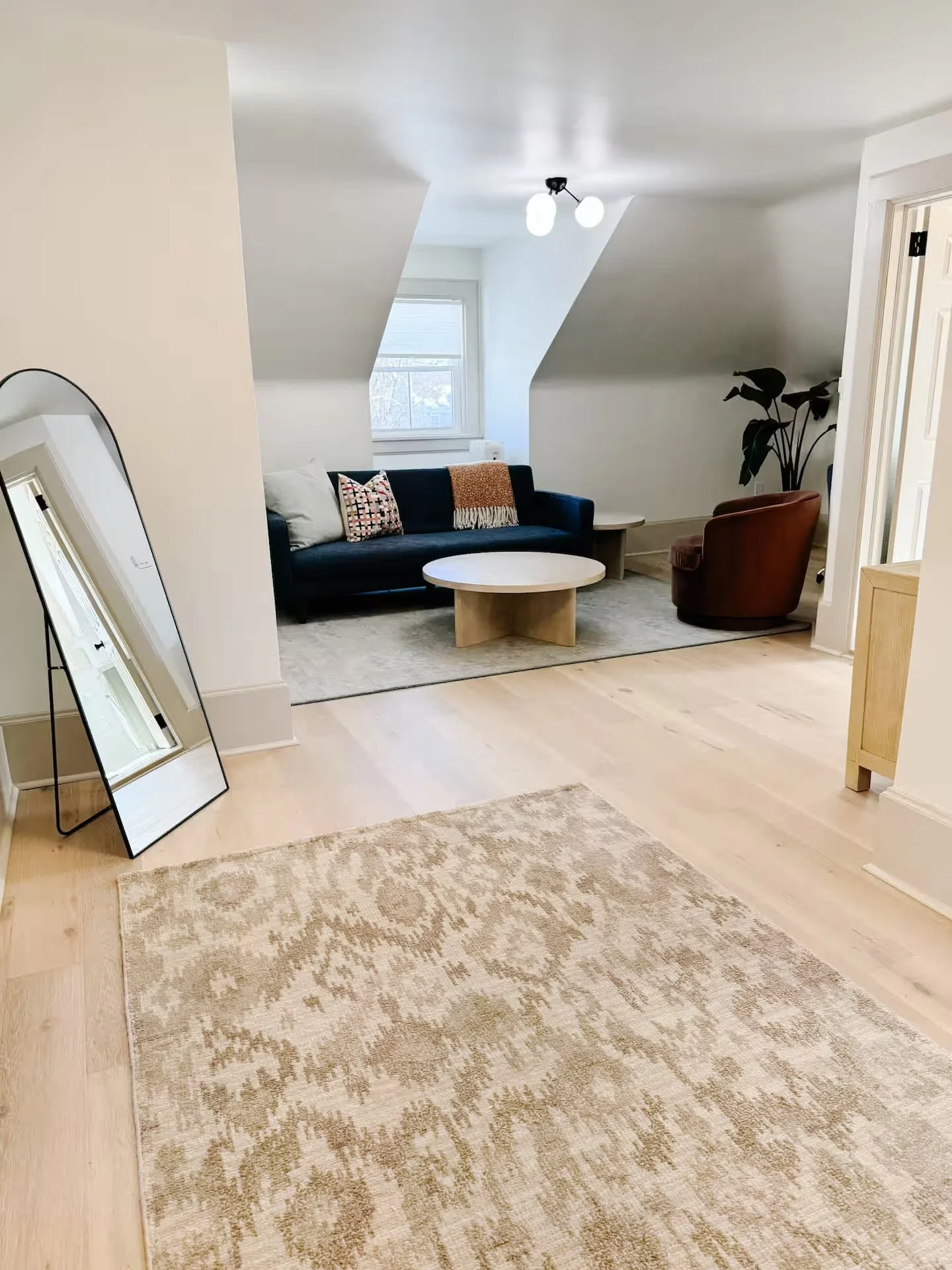 A cozy living room with a beige patterned rug on light wood flooring, a large mirror leaning against a wall, and a dark sofa with accent pillows. There is a round coffee table, a brown armchair, and a tall plant in a corner. A window with white blinds lets in natural light, and a modern ceiling light fixture is visible.