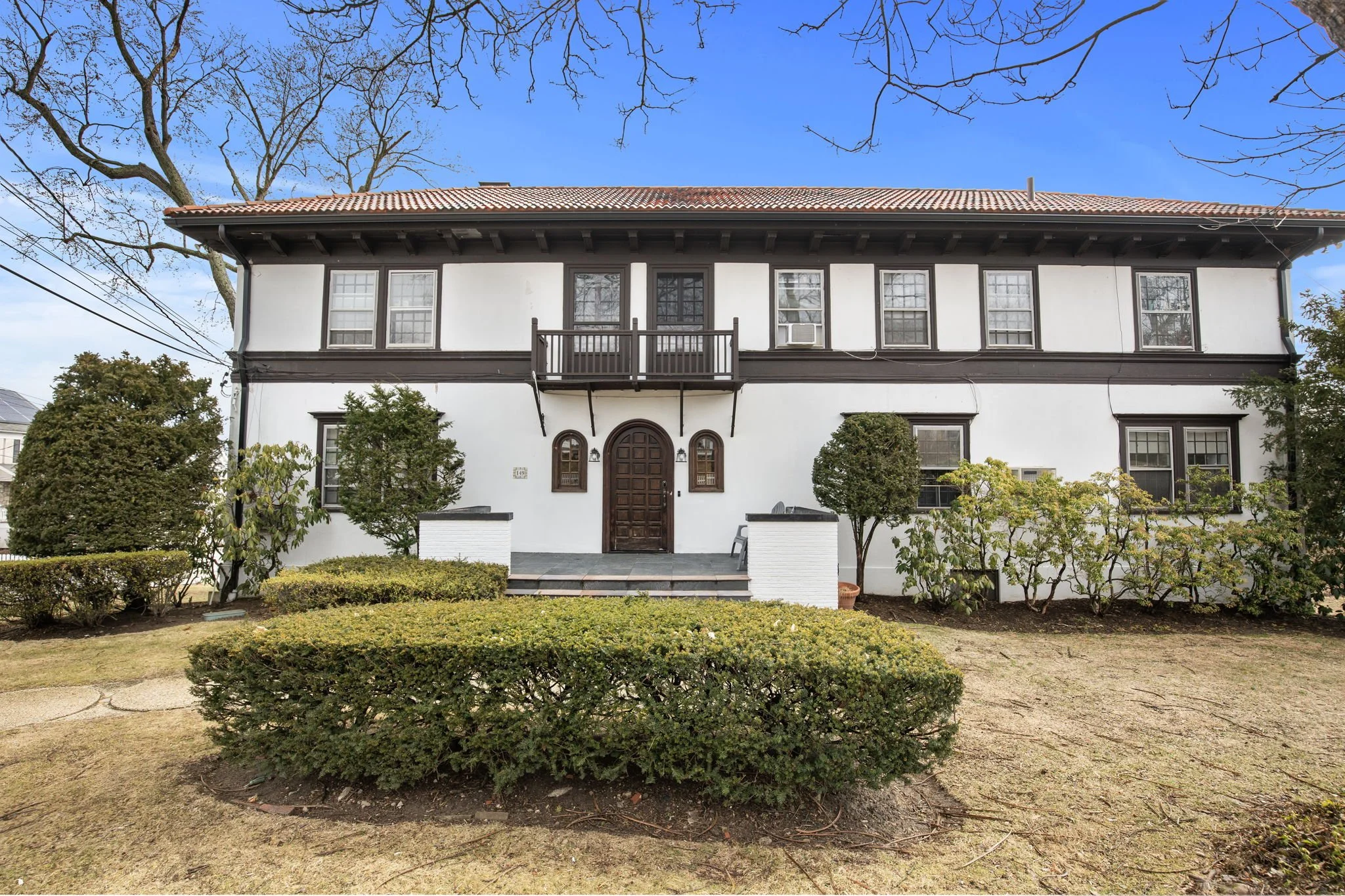 Front view of a two-story white house with dark brown trim, multiple windows, a small balcony, and a black front door; landscaped yard with bushes and small trees, clear blue sky in the background.