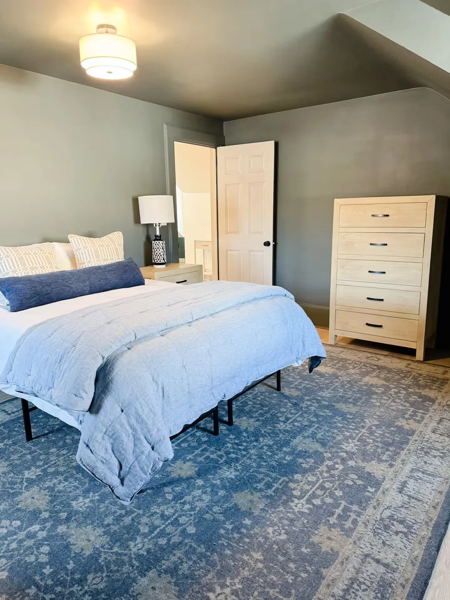 A bedroom with a bed with white and gray bedding, a white nightstand with a lamp, a wooden chest of drawers, and an open closet door. The room has a ceiling light and a blue patterned rug on the floor.
