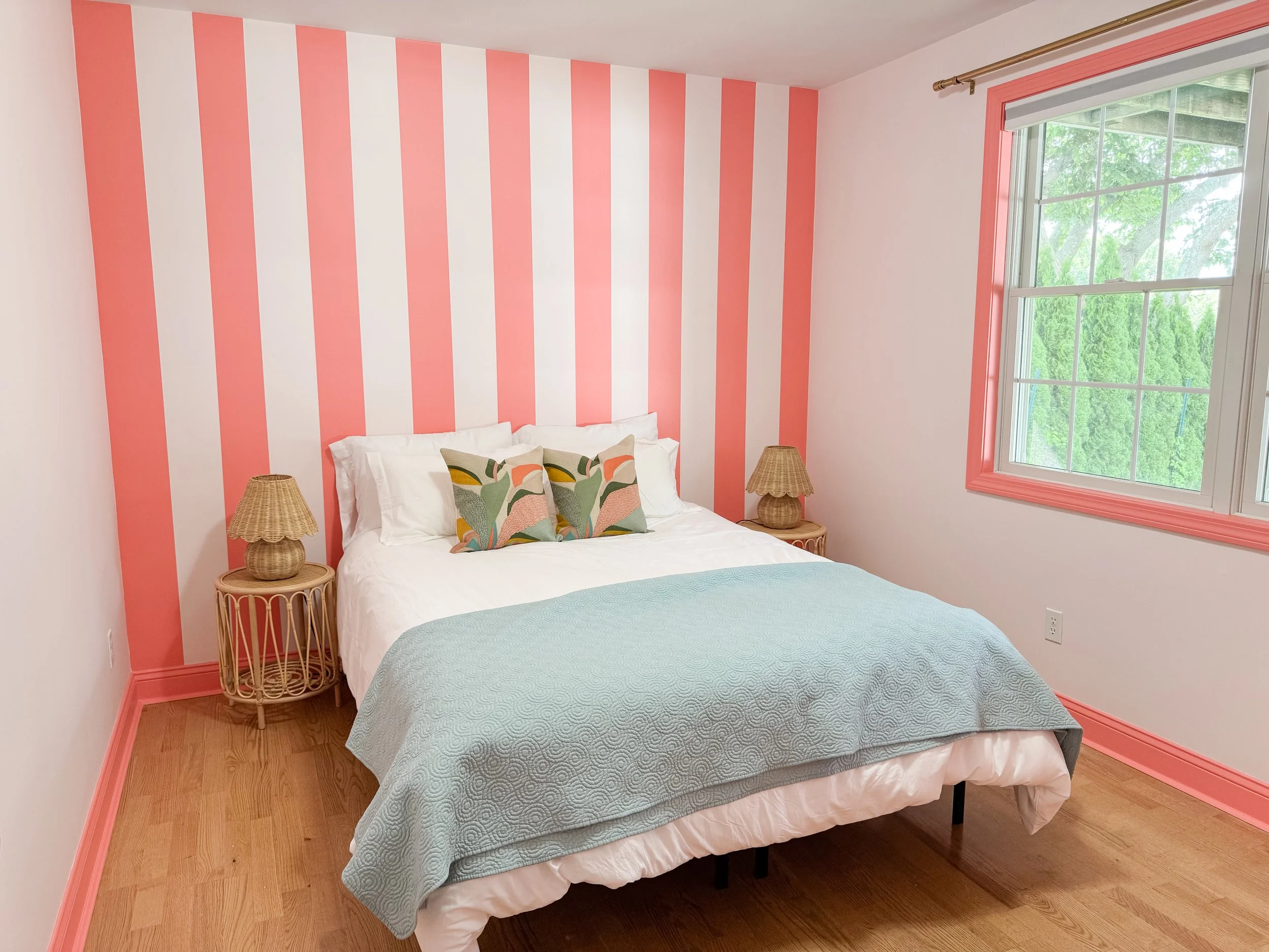 A bedroom with pink and white striped accent wall, wood nightstands with wicker lamps, a bed with white bedding, decorative pillows, a light blue quilt, and a large window with pink trim showing greenery outside.