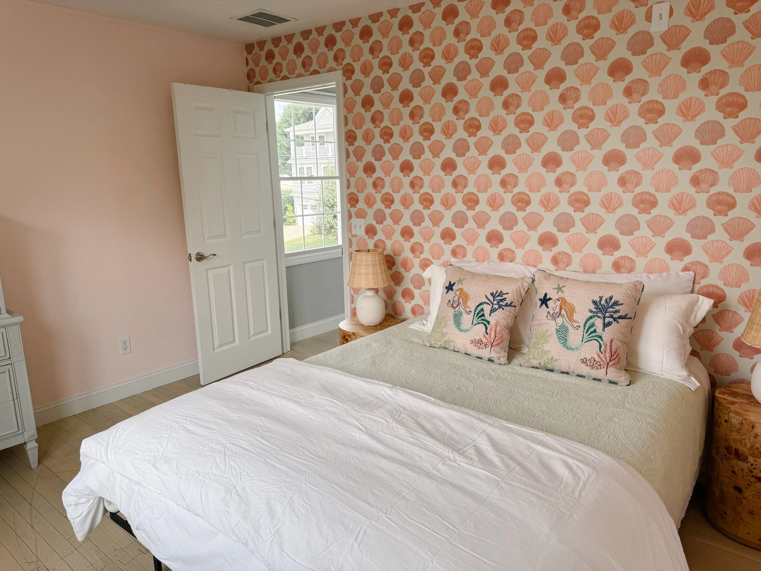 A bedroom with a bed, including pillows with mermaid and seaweed embroidery, a wooden nightstand with a lamp, an open door revealing a window and outdoor greenery, and wallpaper decorated with pink seashells, creating a beach-themed decor.