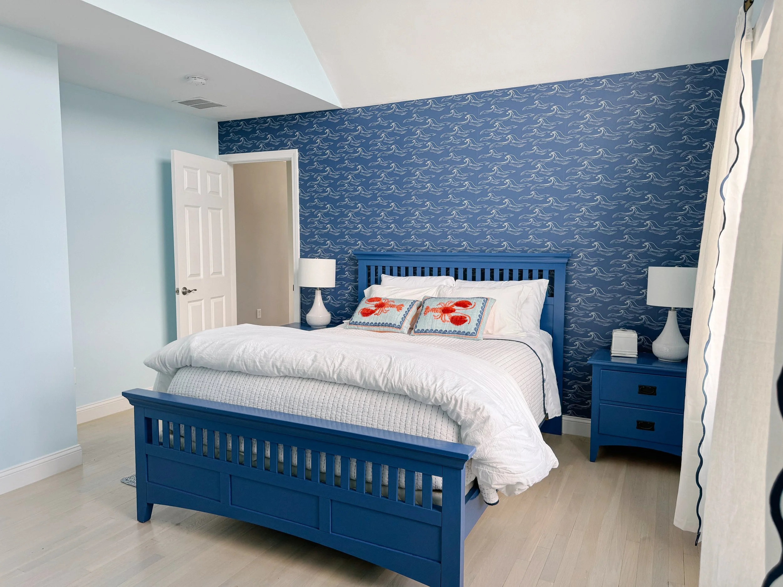 A bedroom with a blue bed frame, white bedding, and decorative pillows with red lobsters, against a blue wall with wave patterns. There are two nightstands with white lamps, a door slightly open, and light-colored wooden floor.