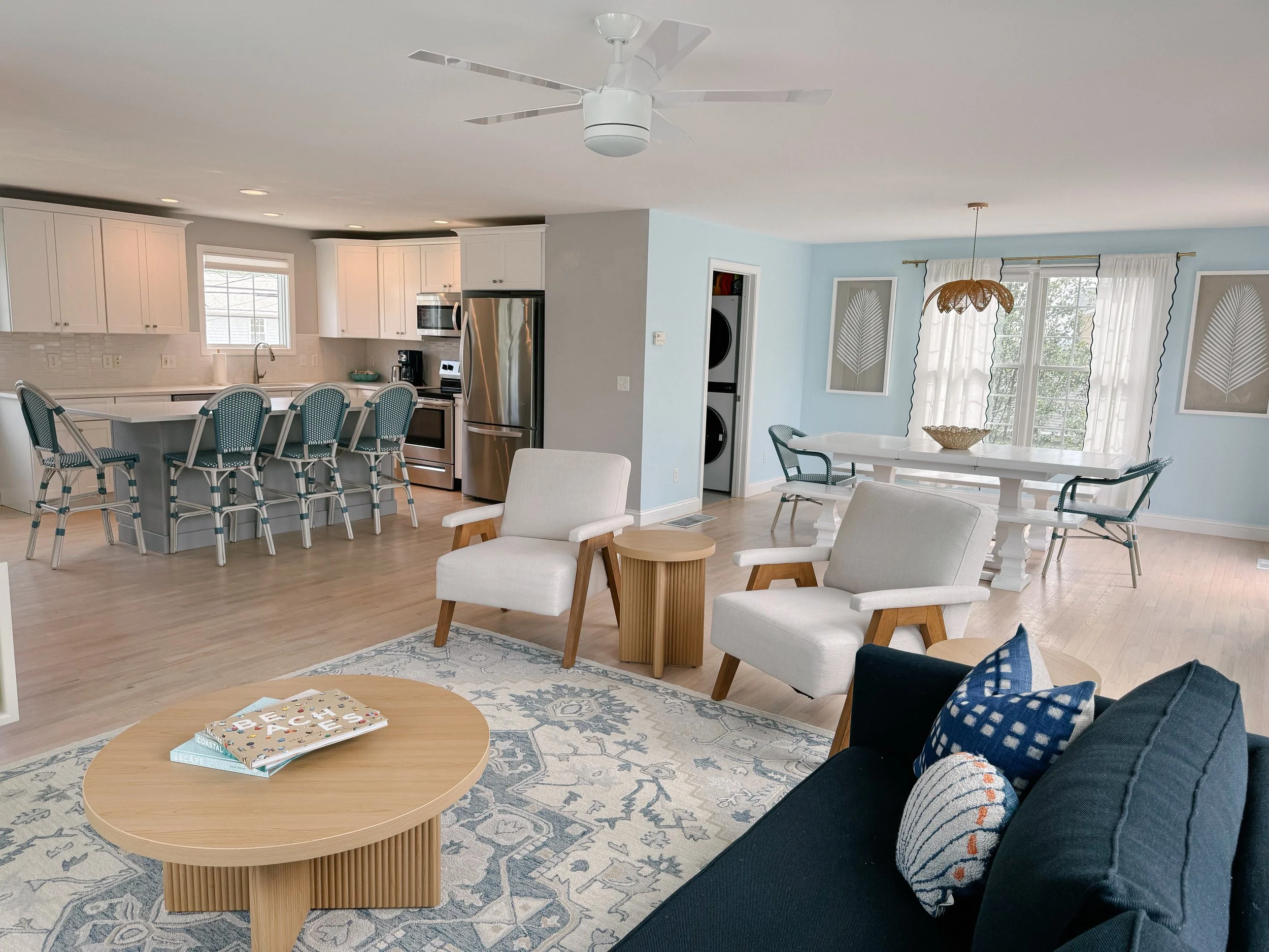 Open-concept living room and kitchen with white cabinets, stainless steel appliances, a breakfast bar with blue and white chairs, a dining table with four chairs, light wood flooring, and large windows with white curtains.