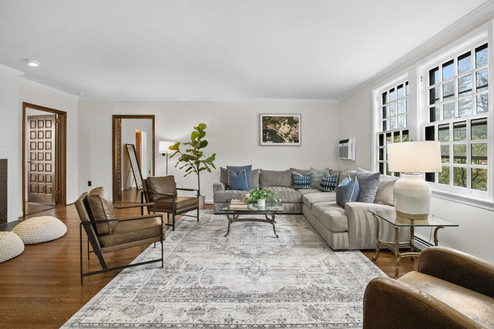 Bright living room with white walls, large windows, beige sectional sofa with blue and patterned pillows, two brown armchairs, a glass coffee table, a large framed landscape painting, a potted plant, and a patterned rug.