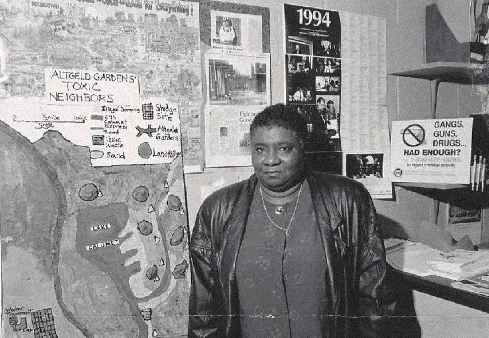A woman with short curly hair wearing a leather jacket and multiple necklaces standing in front of a wall with posters, maps, and signs about local issues. To her right, there's a sign with the text 'Gangs, Guns, Drugs... Had Enough?' and a map of Altgeld Gardens neighborhood.