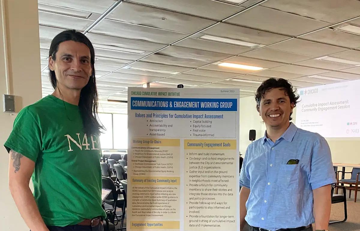 Two men standing indoors near an informational poster about a community engagement working group. One man is wearing a green t-shirt and the other a blue button-up shirt, both smiling at the camera.