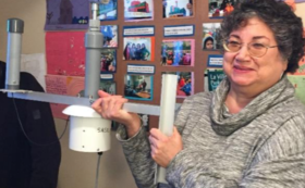 A woman with curly hair and glasses holding a camera stabilizer in an indoor setting, with a colorful wall and photos in the background.