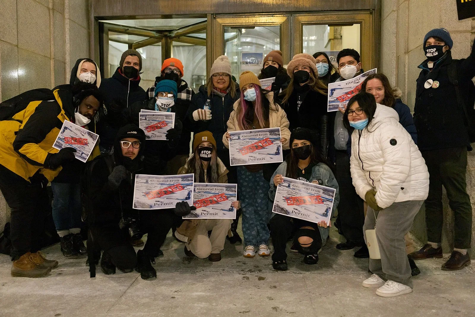 A group of people protesting at a building entrance, many holding signs that say "General Iron Permit" with a red "DENIED" stamp. Some individuals are wearing masks and winter clothing.