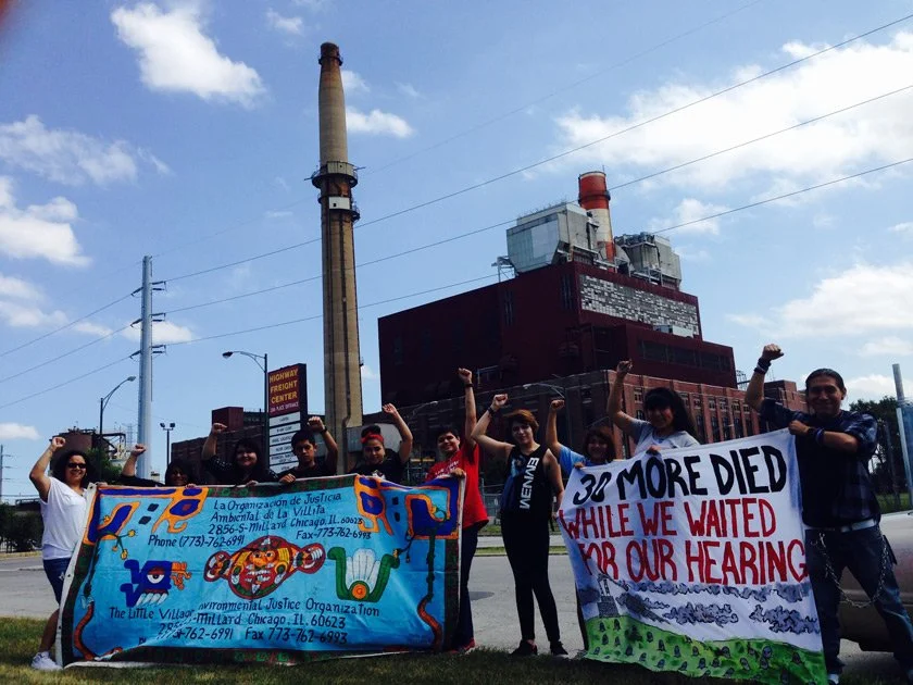 A group of people holding signs and banners protesting environmental issues, with industrial factories in the background.