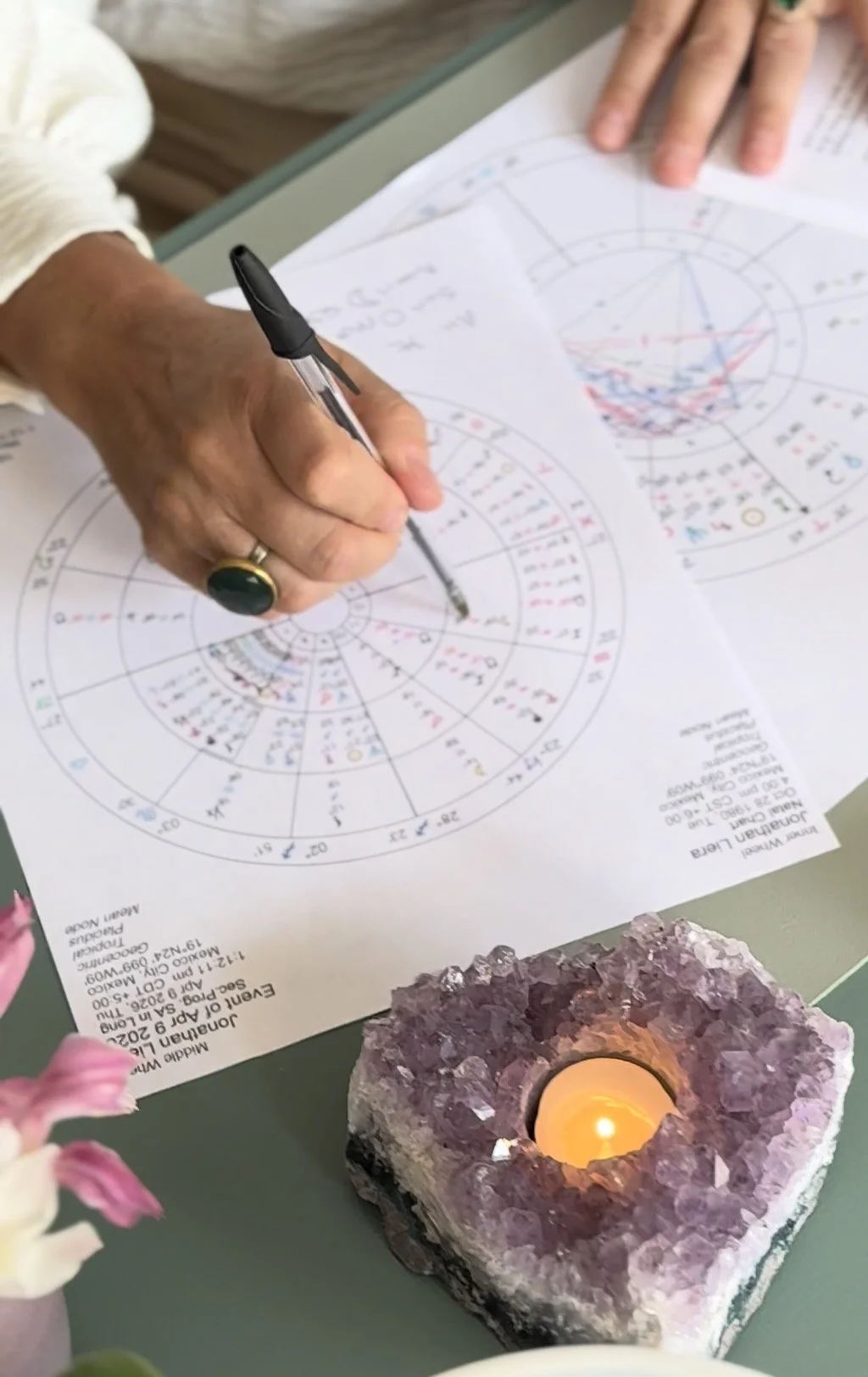 Astrology chart reading. A person writes on a detailed astrological birth chart with a black pen. There is a lit candle in a purple geode holder on the table and some pink flowers nearby.