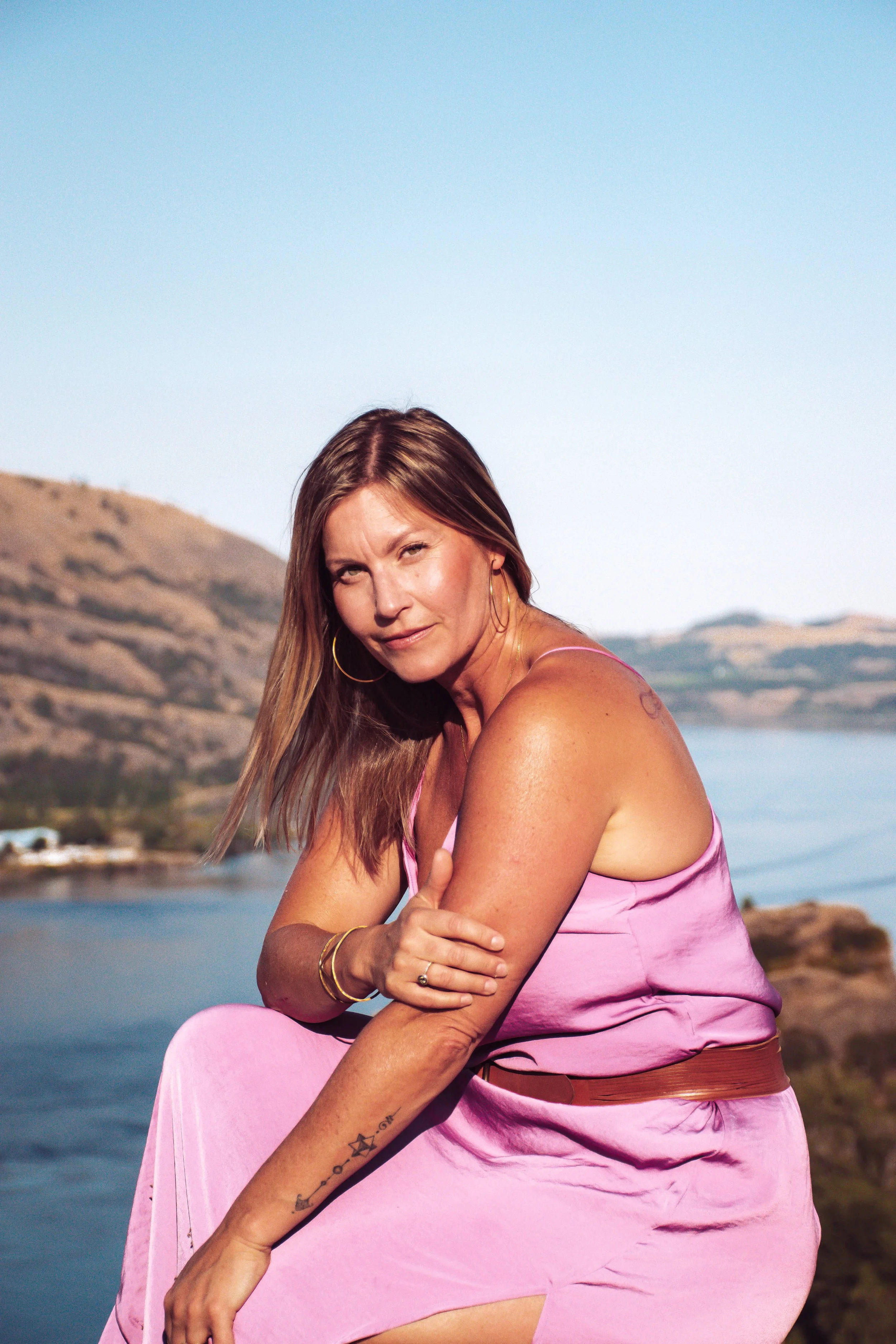 Tara Jade Nichols Spiritual Mentorship. A woman in a pink dress sitting on rocks near a body of water, with hills in the background and clear skies.