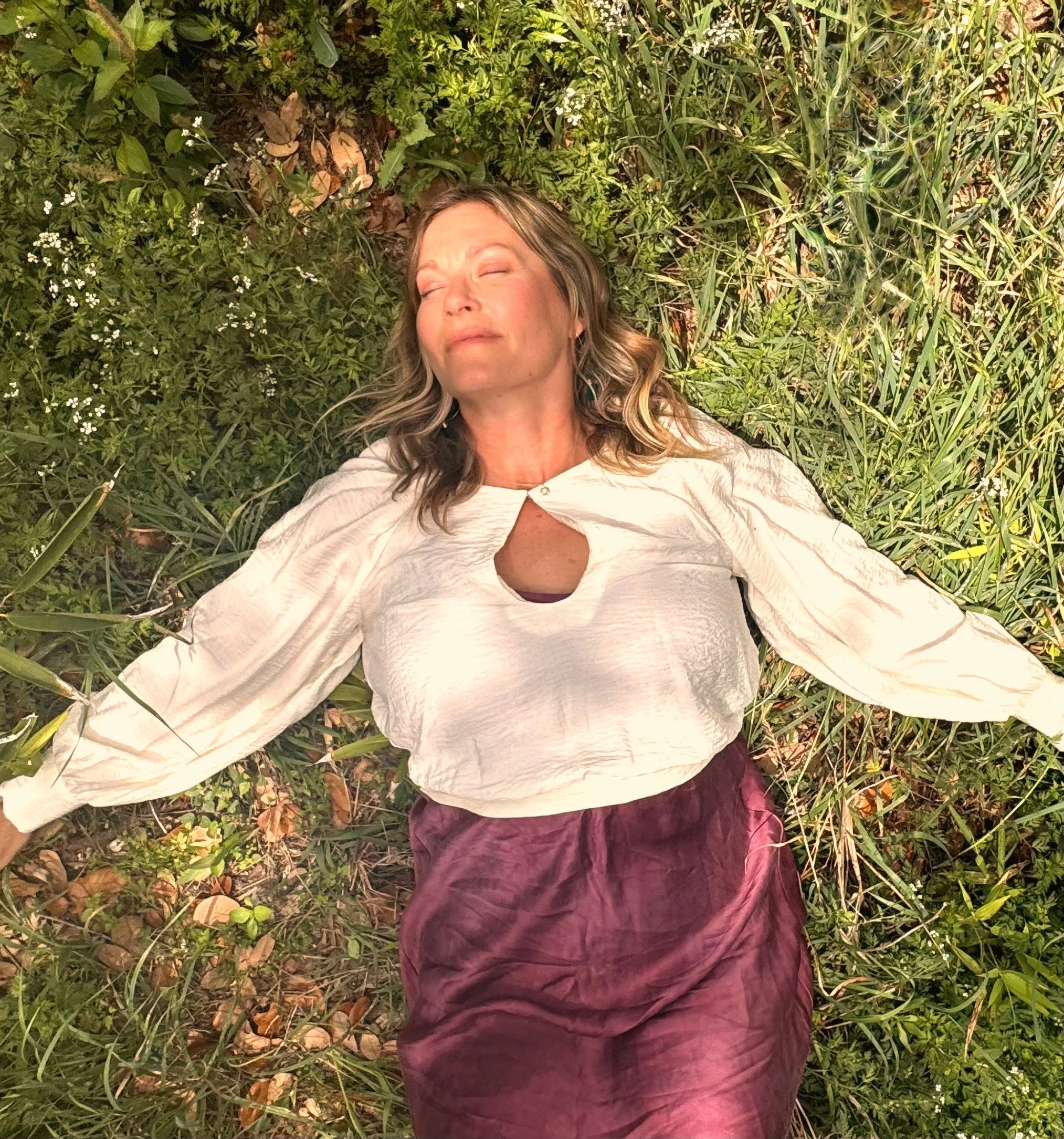 A woman with curly hair and light skin lying on her side on grass, with eyes closed and a peaceful expression, surrounded by green plants and small white flowers.